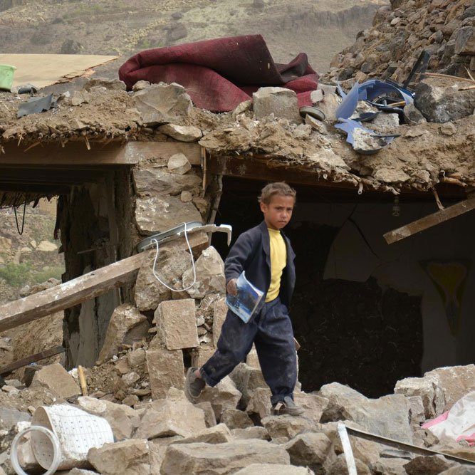 L'image montre un enfant marchant à travers des décombres d'un bâtiment détruit. Les murs sont effondrés et de nombreux débris, tels que des pierres et des objets éparpillés, sont visibles autour de lui. L'environnement semble être celui d'une zone touchée par un désastre, avec un paysage en arrière-plan qui pourrait suggérer des conditions difficiles. L'enfant porte des vêtements sombres, et son expression évoque une certaine gravité face à la situation environnante.