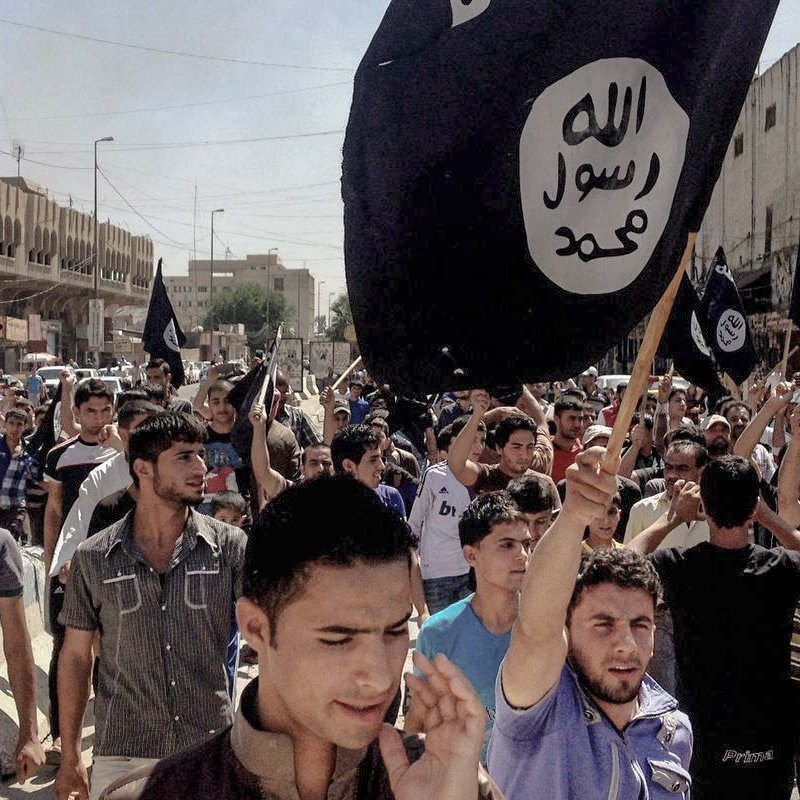 L'image montre une foule de personnes rassemblées dans une rue, tenant des drapeaux noirs emblématiques. Les manifestants semblent mobilisés et expriment une forte émotion. L'environnement urbain autour d'eux montre des bâtiments et une rue animée, suggérant un événement significatif ou une manifestation.