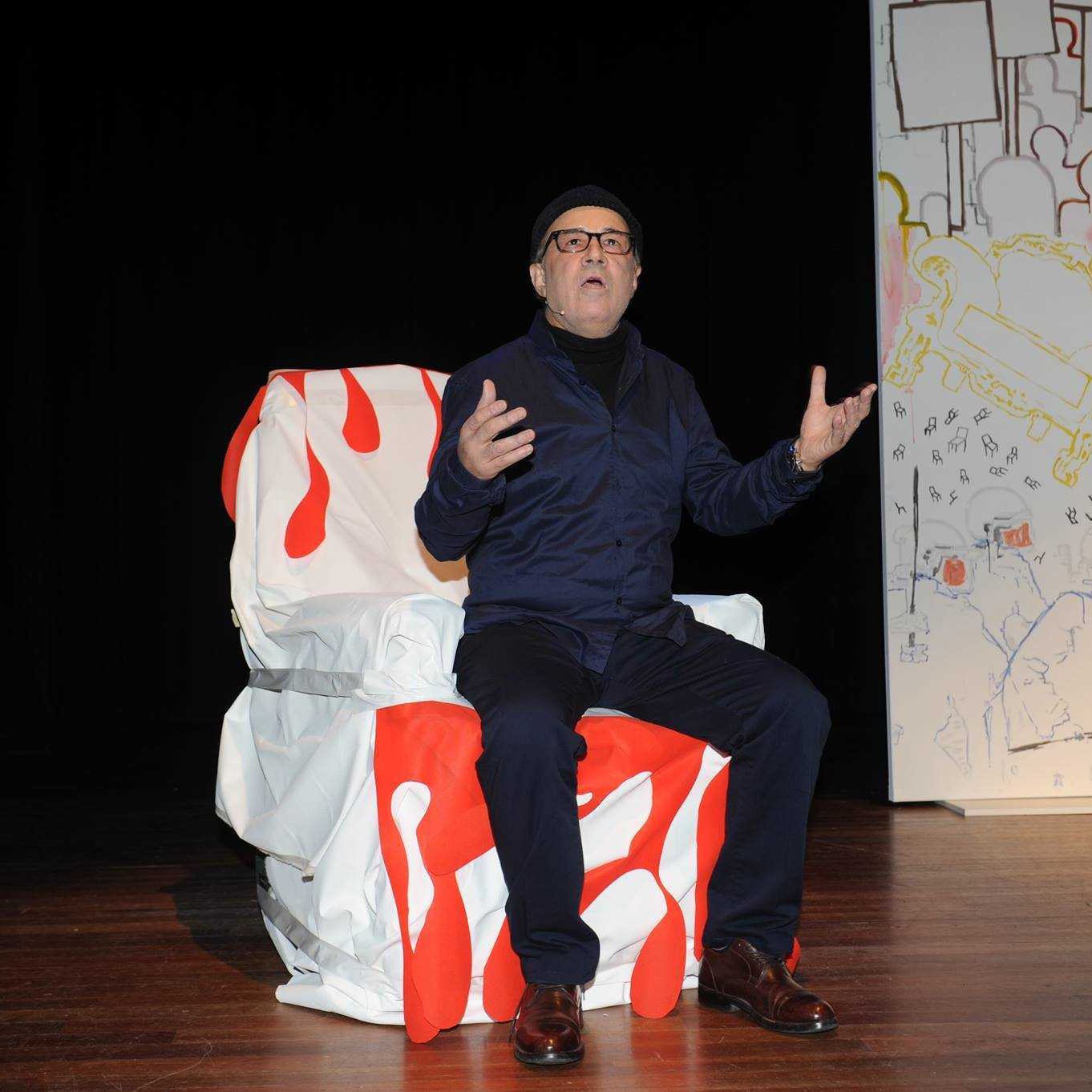 The image features a man sitting on a uniquely designed chair that has a colorful, artistic pattern, predominantly featuring red and white. He is wearing a dark outfit, including a jacket and glasses, and appears to be expressing himself with his hands raised, as if making a point during a performance or presentation. The background includes a large, stylized backdrop with a simple, colorful design. The setting seems to be a stage, suggesting a theatrical or artistic context.