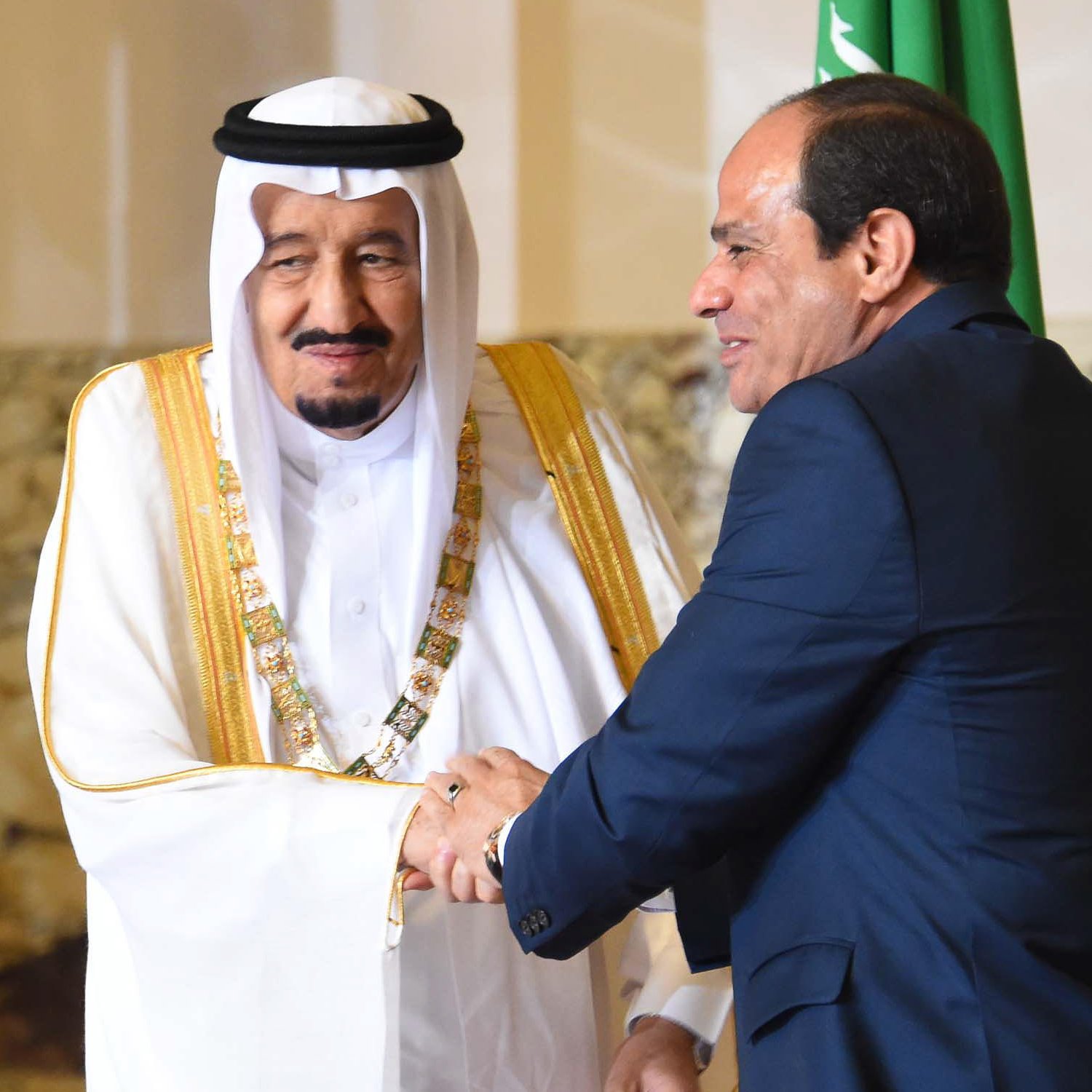 L'image montre deux hommes en costume, se serrant la main dans un cadre formel. L'un est vêtu d'une robe traditionnelle saoudienne et d'un couffin, tandis que l'autre porte un costume sombre. Ils semblent échanger un sourire, ce qui indique une interaction amicale. En arrière-plan, on peut apercevoir des décorations et des drapeaux, suggérant un contexte diplomatique ou officiel.