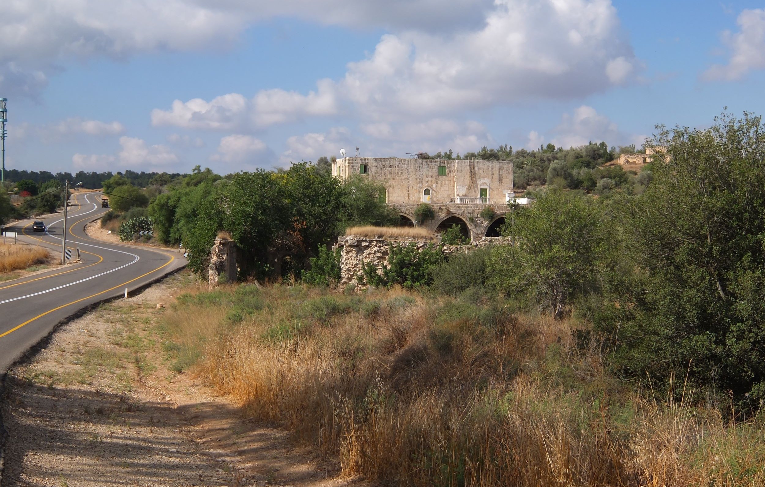 L'immagine mostra un paesaggio rurale caratterizzato da una strada curvilinea che si snoda tra la vegetazione. Ai lati della strada, ci sono prati e aree erbose, con un'atmosfera tranquilla e naturale. Sullo sfondo, spicca una vecchia costruzione in pietra, apparentemente in rovina, con archi visibili, circondata da alberi e vegetazione. Nel cielo, si possono vedere alcune nuvole sparse, che completano il quadro di un ambiente sereno e suggestivo.