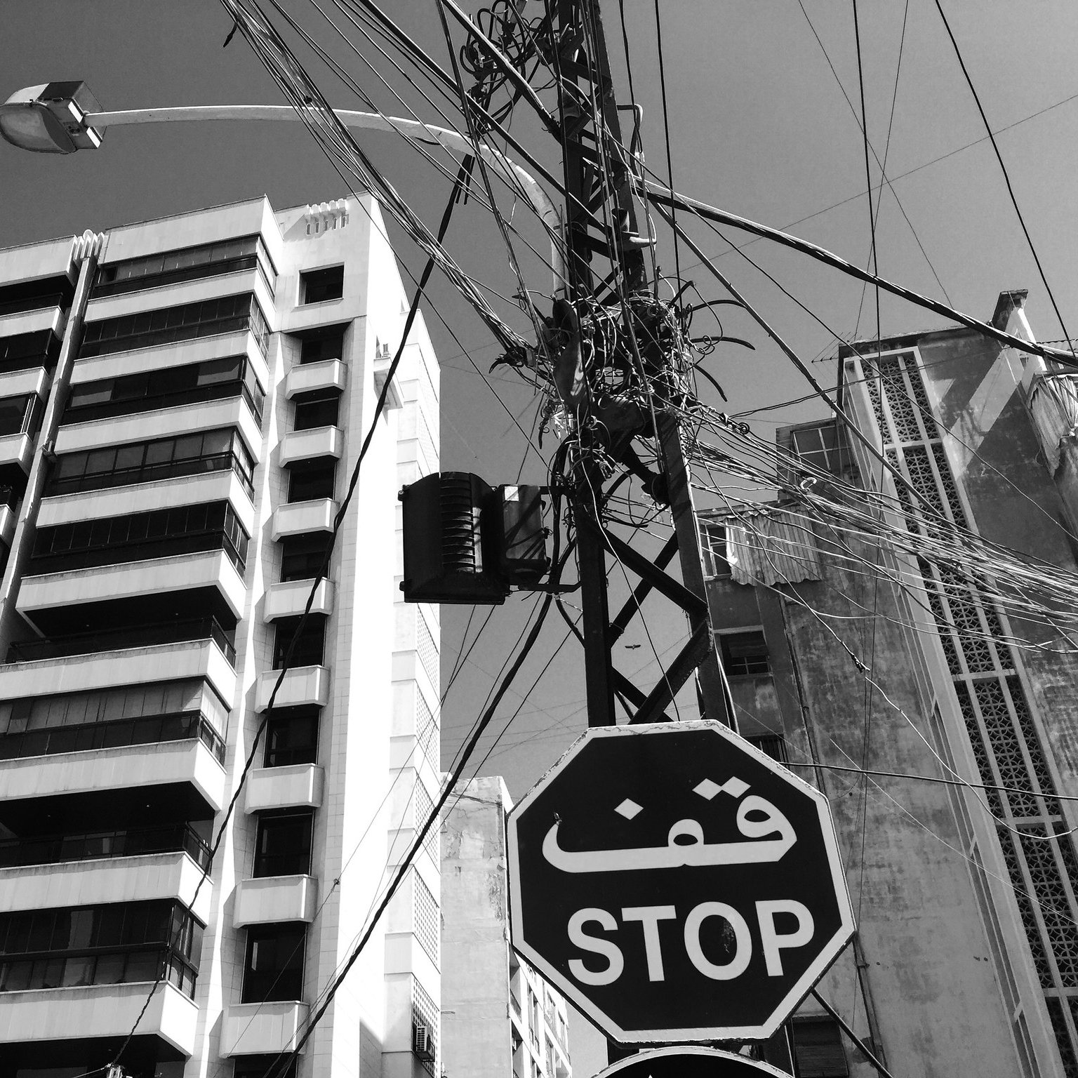 L'image montre une scène urbaine monochrome. À gauche, on aperçoit un bâtiment moderne avec plusieurs étages et balcons. À droite, un poteau électrique est visible, avec des fils enchevêtrés qui s'étendent dans le ciel. Il y a un panneau de signalisation en forme de hexagone avec le mot "STOP" écrit en arabe, ajouté à l'ambiance citadine. Le ciel est clair, ce qui donne une impression de luminosité à l'ensemble de la scène.