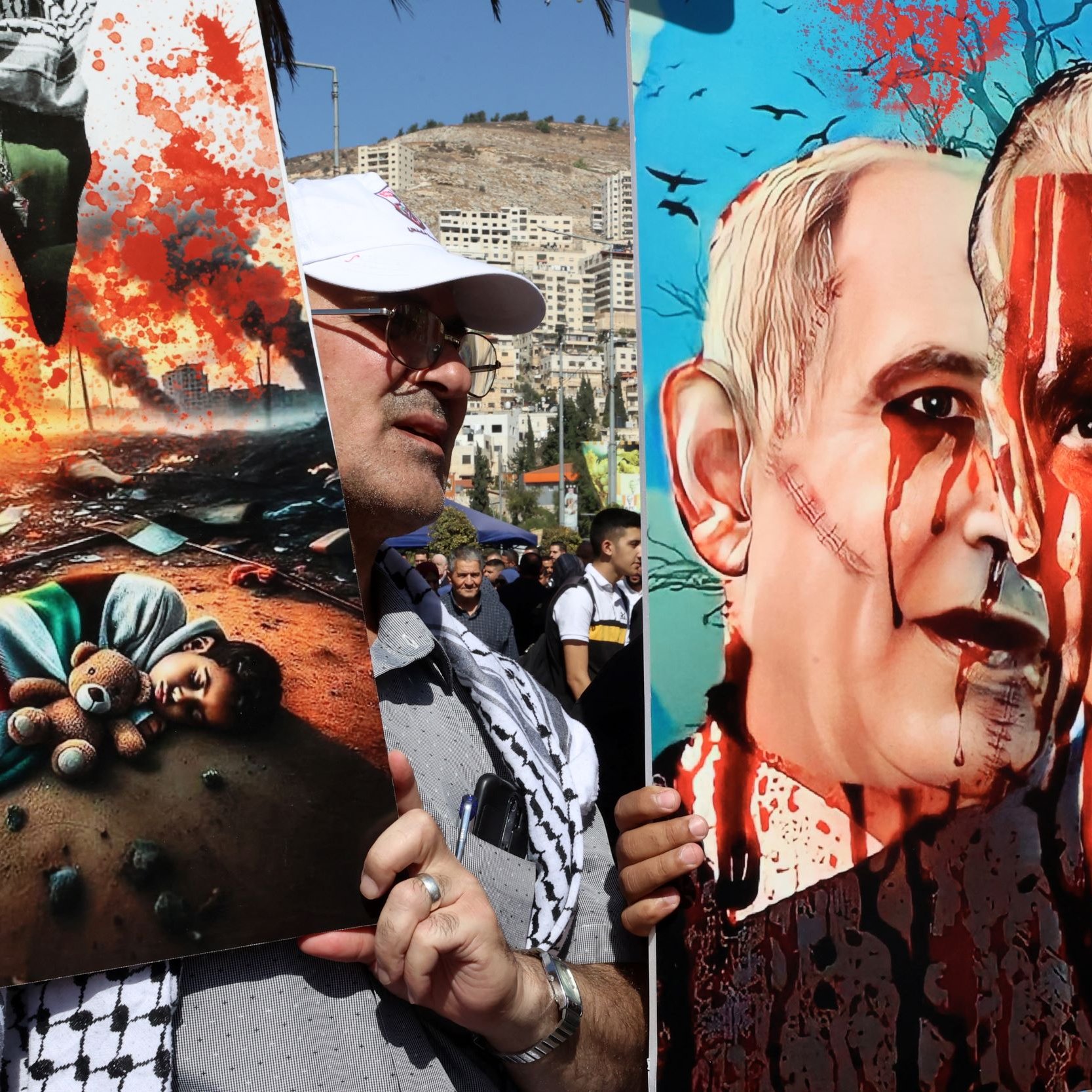 The image features two striking pieces of protest art held by a person. On the left, there is a depiction of a devastated scene showing a child's lifeless body and a teddy bear amidst destruction, conveying a powerful message about the impact of conflict on innocent lives. On the right, another artwork portrays two figures resembling political leaders, with exaggerated features and blood splatters, suggesting themes of violence and political aggression. The overall mood of the image is one of protest and strong emotional expression regarding war and its consequences.