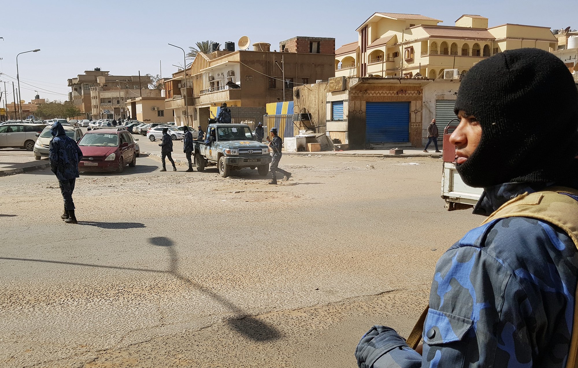 L'image montre une scène urbaine où des policiers armés sont présents dans une rue. On peut voir plusieurs membres des forces de l'ordre, certains portant des uniformes camouflés et des masques, tout en étant attentifs à leur environnement. Il y a des véhicules de police, ainsi que des voitures stationnées à proximité. En arrière-plan, on aperçoit des bâtiments de style architectural varié. L'atmosphère semble tendue, évoquant un contexte de sécurité renforcée.