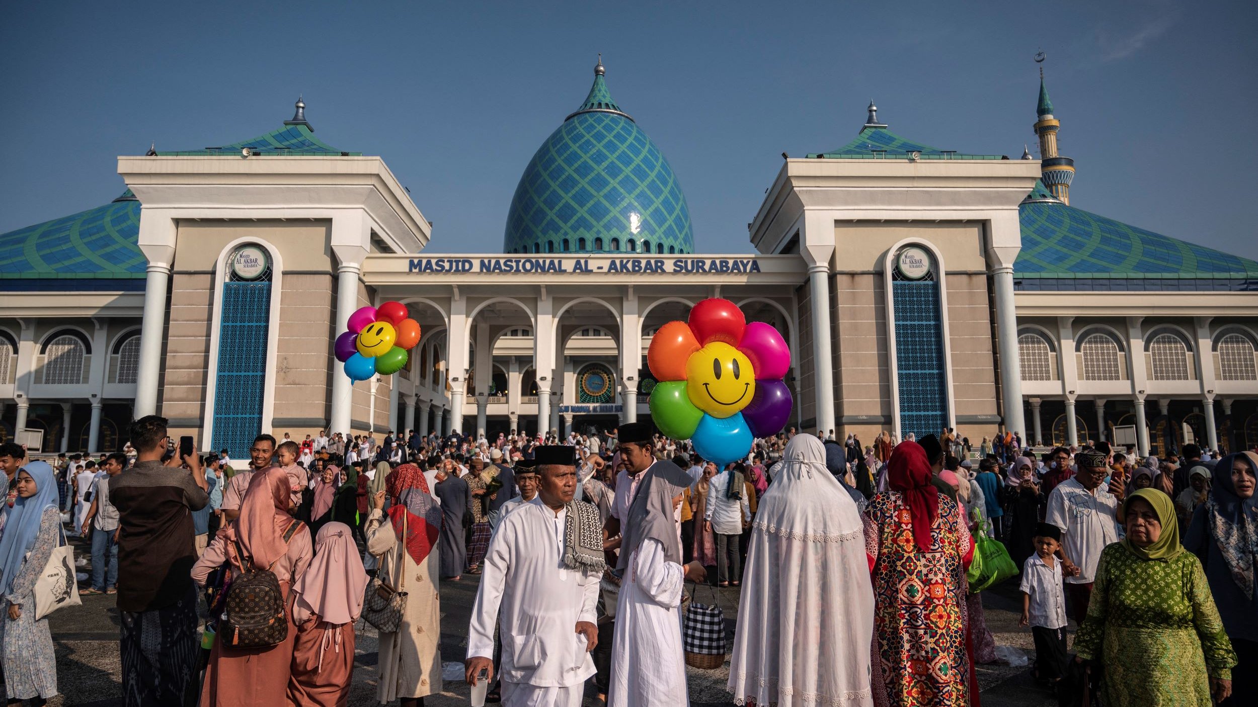 L'image montre un grand rassemblement devant la Masjid Nasional Al-Akbar de Surabaya, en Indonésie. La mosquée est impressionnante, avec des dômes bleu turquoise et une architecture moderne. Beaucoup de personnes sont présentes, certaines portant des vêtements traditionnels, et elles semblent participer à une célébration ou un événement. Des ballons colorés en forme de visages souriants ajoutent une touche festive à la scène. Le ciel est clair, suggérant une belle journée ensoleillée.