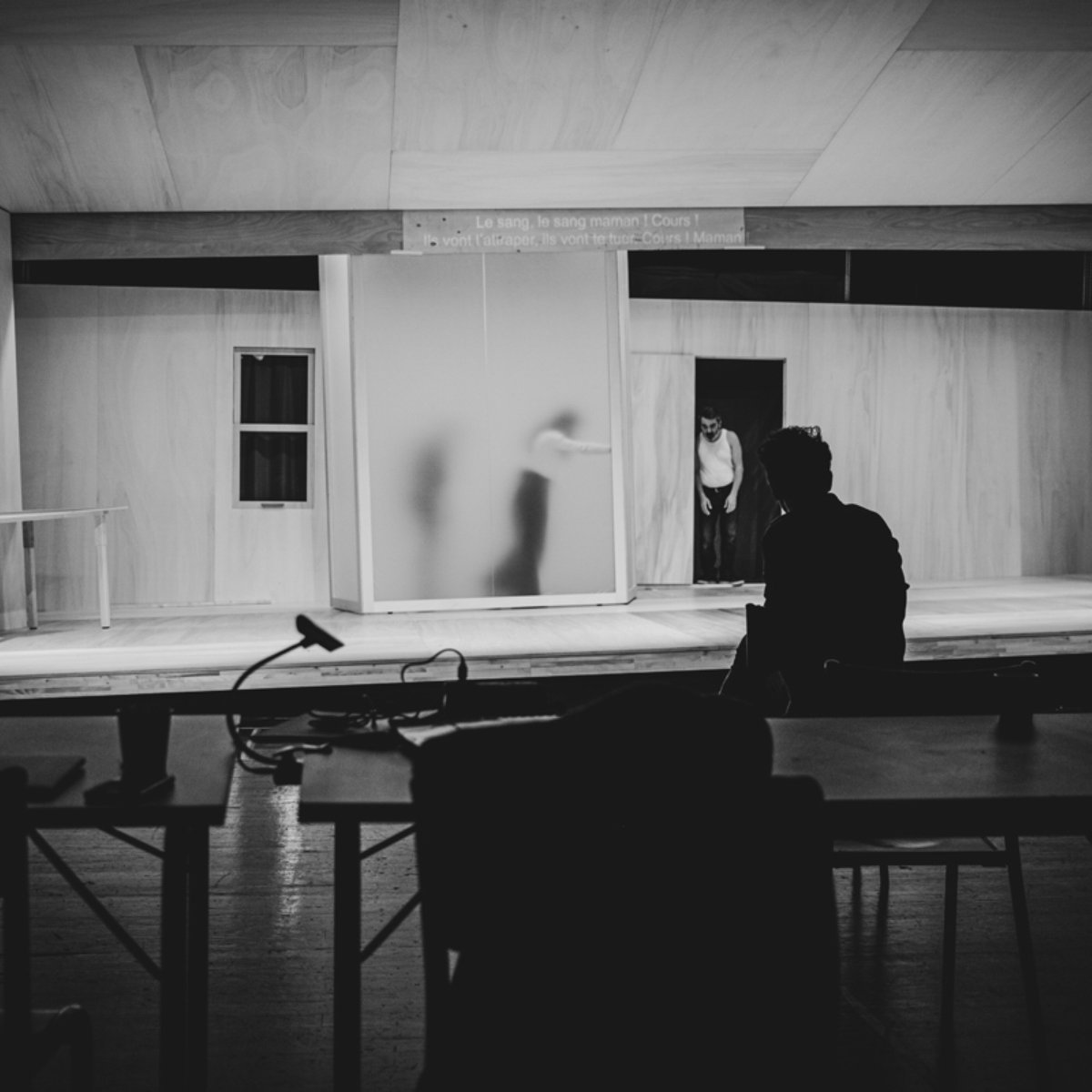 L'image montre un espace scénique avec des murs en bois clair et un éclairage tamisé. On peut apercevoir une silhouette floue à l'arrière-plan, probablement en train de se déplacer. Au premier plan, une personne est assise de dos à une table, observant la scène. L'ambiance est sombre et intimiste, suggérant un moment d'attente ou de concentration avant une représentation. Les éléments de décor, comme les fenêtres et un meuble, ajoutent à l'atmosphère théâtrale.