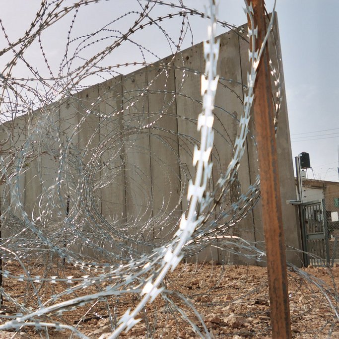 L'image montre un paysage marqué par des barbelés disposés en spirale et un mur en béton en arrière-plan. Le terrain est rocheux et dépouillé, ce qui contribue à une ambiance austère et sécurisée. Les barbelés créent une impression de barricade ou de clôture, suggérant une séparation ou un isolement. Le ciel au-dessus est clair, ajoutant un contraste avec les éléments de sécurité présents dans le paysage.