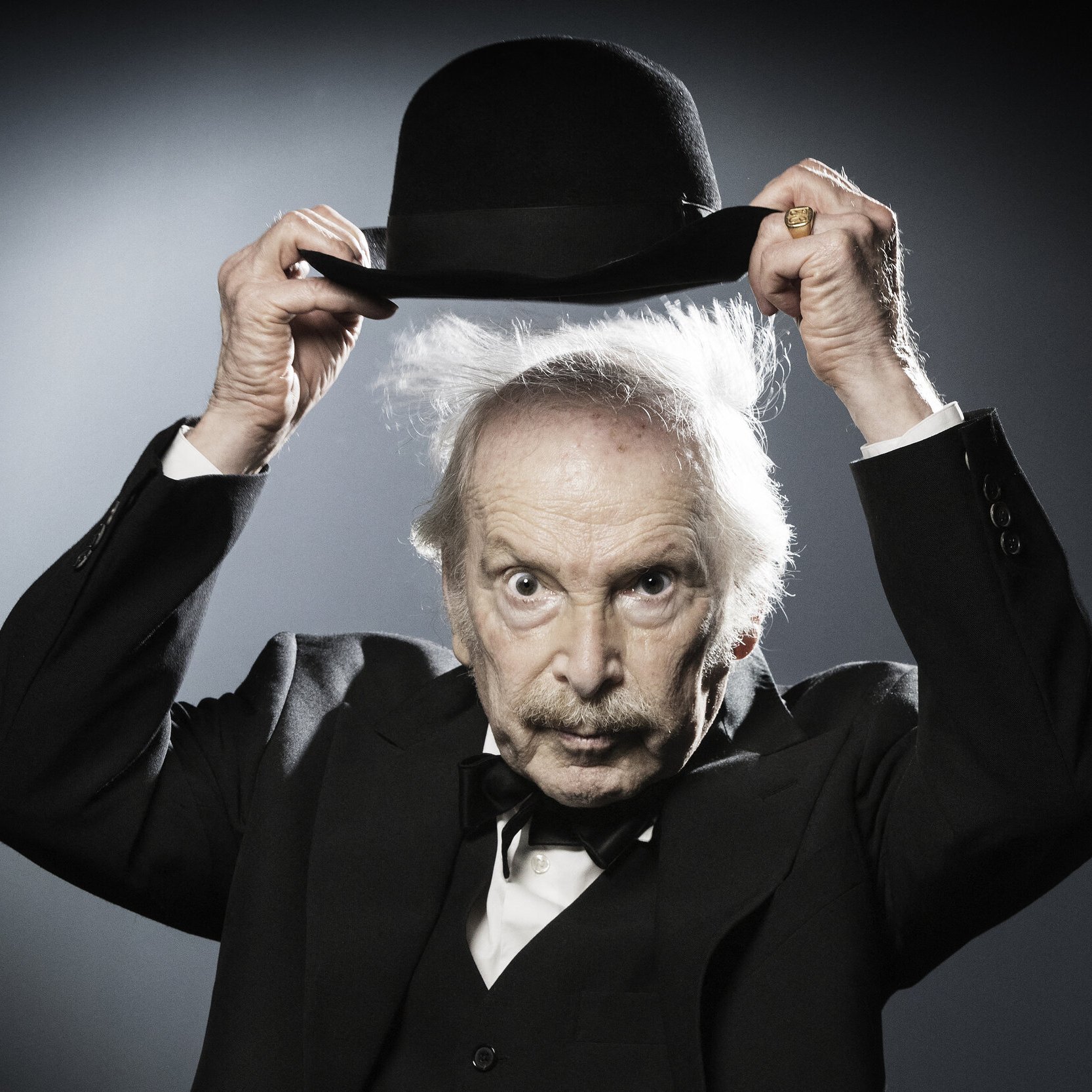 L'image montre un homme âgé portant un costume noir avec un nœud papillon. Il tient un chapeau melon avec une main, et son autre main semble légèrement levée. Ses cheveux sont ébouriffés, ce qui donne un aspect excentrique à son expression. L'arrière-plan est sobre, avec une lumière qui crée un effet dramatique, mettant en valeur son visage et son chapeau.