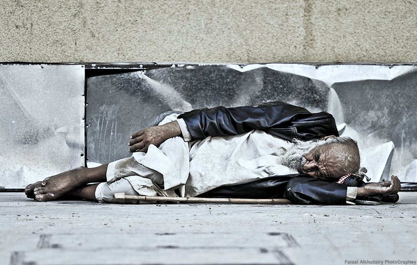 L'image montre un homme âgé allongé sur le sol, semblant endormi. Il porte des vêtements usés et semble être dans une position de repos sur un trottoir. Son visage est marqué par le temps et il est entouré d'un environnement urbain. L'atmosphère évoque une certaine mélancolie et une réflexion sur la vie des personnes sans abri.