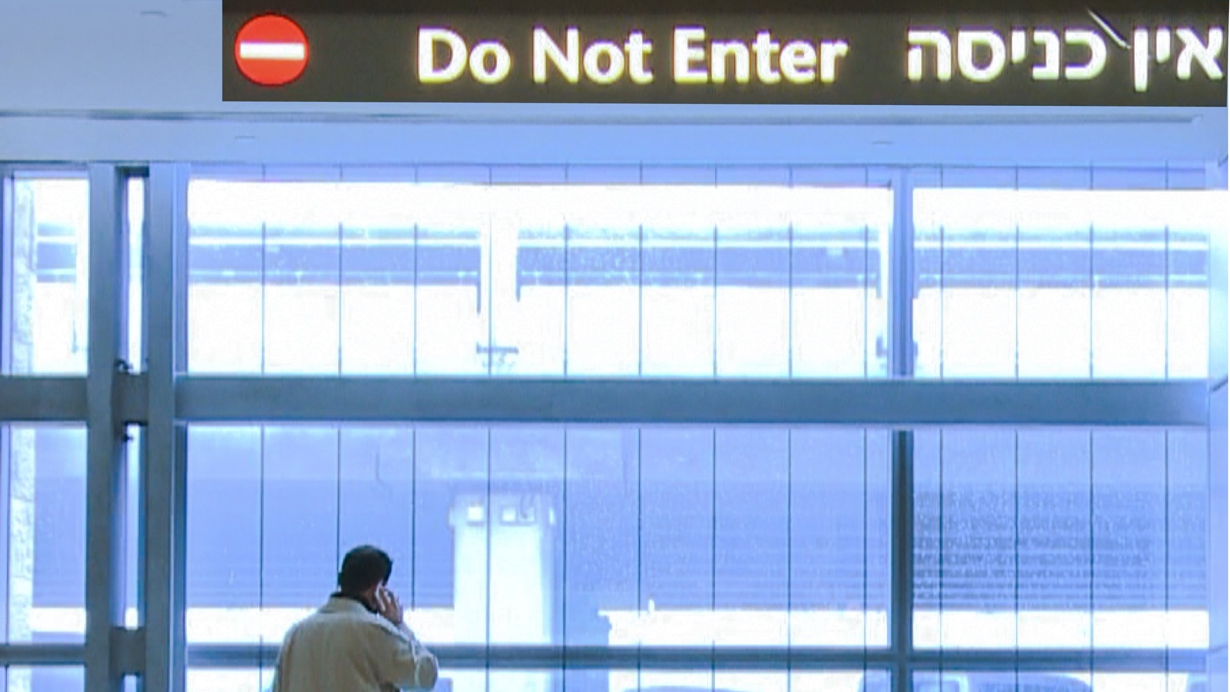 L'image montre un homme debout près d'une grande fenêtre dans un espace intérieur moderne. Il porte un long manteau clair et semble être en train de parler au téléphone. Au-dessus de lui, il y a une signalisation en trois langues, indiquant "Do Not Enter" en anglais et une phrase en hébreu, ainsi qu'un panneau rouge avec un symbole d'interdiction. L'espace est bien éclairé, avec des murs en verre et une ambiance assez minimaliste.