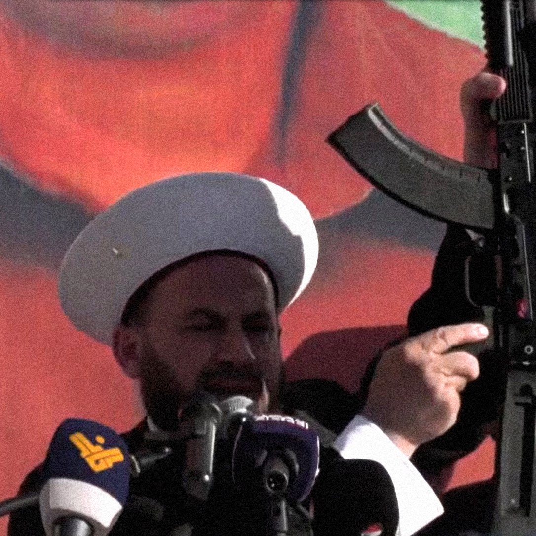 L'image montre deux hommes portant des chapeaux traditionnels, se tenant devant un grand panneau ou une affiche représentant une figure politique ou historique. L'un des hommes brandit une arme, probablement un fusil d'assaut, en faisant un geste qui attire l'attention du public. L'atmosphère semble solennelle et les deux hommes semblent s'adresser à une foule, ce qui suggère un contexte d'engagement politique ou religieux.