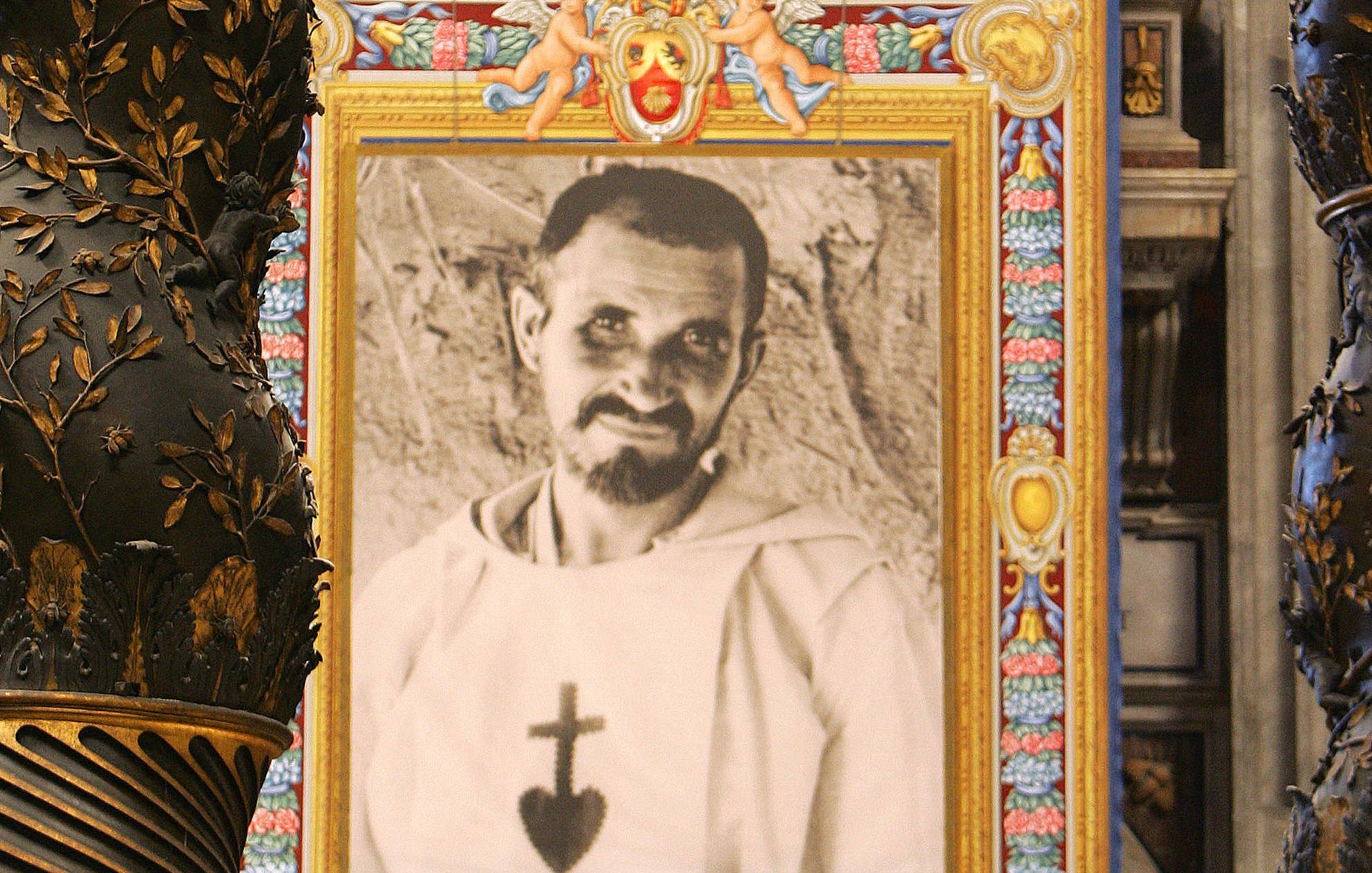 L'image montre un intérieur majestueux, probablement dans une église ou une cathédrale, avec des colonnes ornées de motifs dorés et des détails complexes. Au centre, il y a un grand portrait en noir et blanc d'un homme, qui semble vêtu d'une robe religieuse, encadré de couleurs vives et de décorations. Ce cadre attire l'œil et contraste avec la richesse des ornements de l'architecture environnante. Les murs et les éléments de la pièce évoquent une atmosphère sacrée et historique.