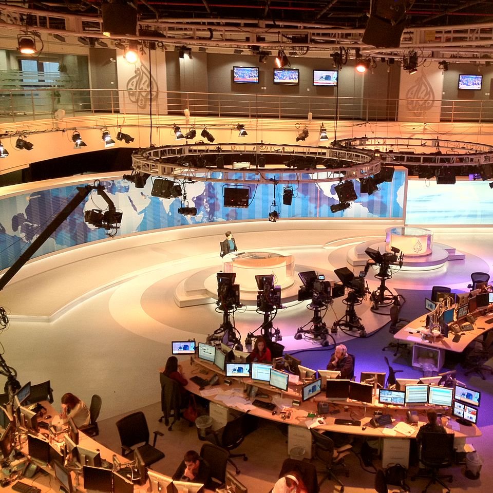 L'image montre un studio de télévision moderne, probablement utilisé pour l'information. On y voit plusieurs caméras placées sur des trépieds, ainsi que des moniteurs et des bureaux avec des ordinateurs. Le décor est élégant, avec des panneaux de fond aux couleurs douces et des éclairages suspendus. Au centre, il y a un espace pour le présentateur, entouré d'une atmosphère professionnelle et technologique. L'ensemble donne une impression de dynamisme et de préparation pour diffuser des nouvelles en direct.