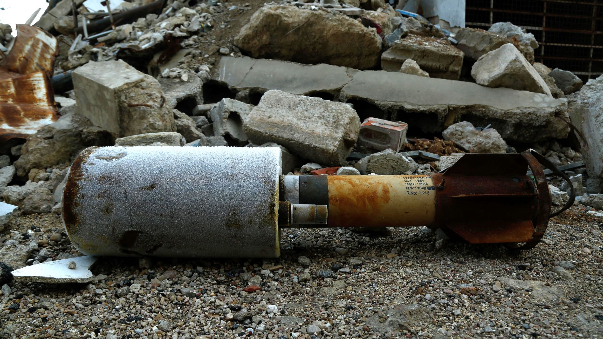La imagen muestra un artefacto explosivo que yace en el suelo entre escombros de una construcción destruida. El objeto está parcialmente cubierto de polvo y parece haber estado en el lugar durante algún tiempo, lo que sugiere que puede estar relacionado con un conflicto o un evento violento. Los escombros a su alrededor incluyen trozos de concreto y metal, lo que refuerza la impresión de un entorno devastado.