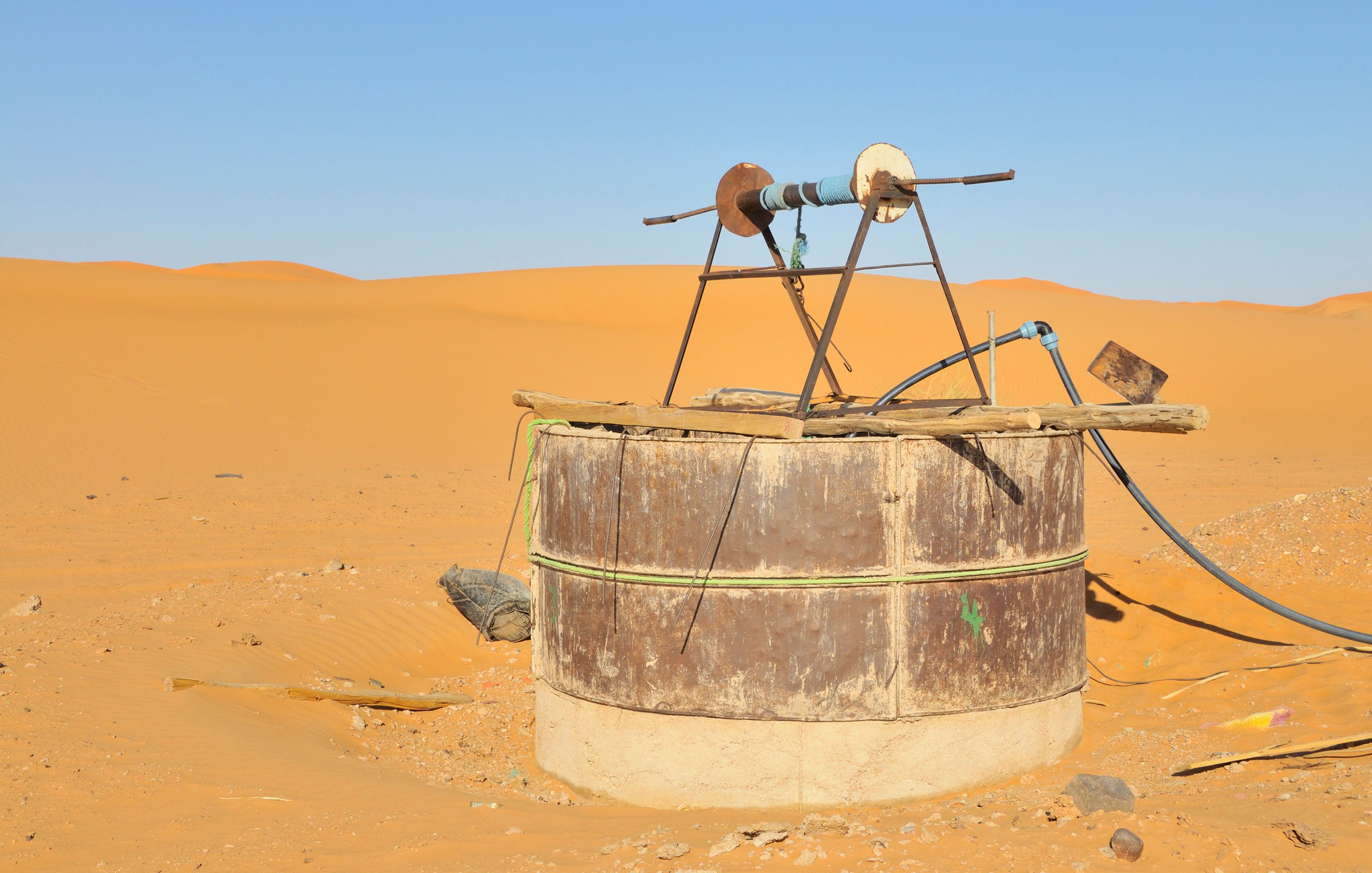 L'image montre un puits situé au milieu d'un désert, entouré de dunes de sable doré. Le puits est constitué d'une structure en métal circulaire, partiellement enterrée dans le sol. Au sommet, il y a un système de poulies qui semble être utilisé pour tirer de l'eau. Le ciel est dégagé, avec une lumière vive qui illumine le paysage désertique. L'environnement est aride et semble isolé, évoquant une atmosphère de tranquillité et de solitude.