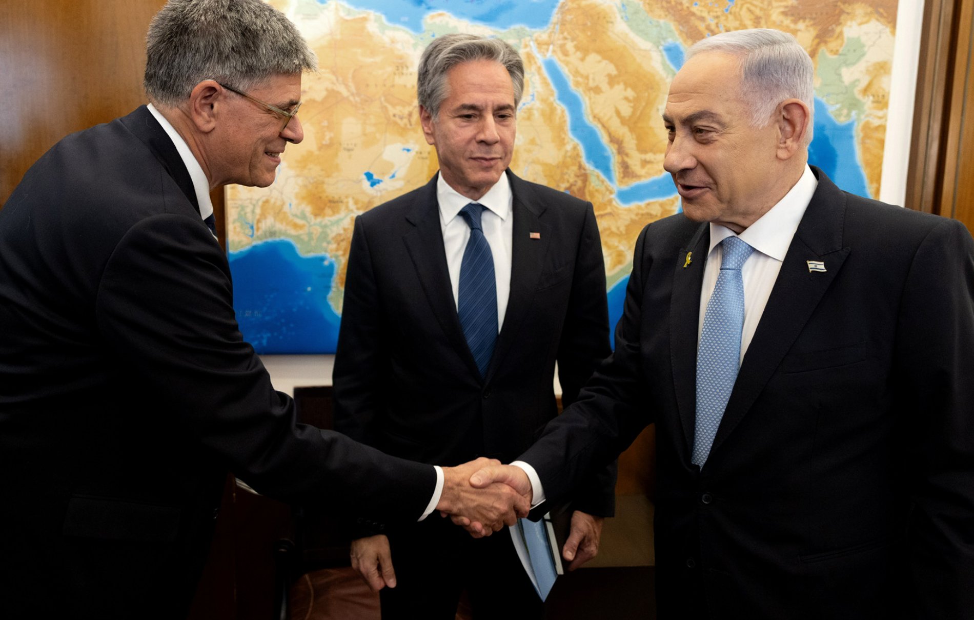 L'image montre trois hommes dans un cadre formel. À gauche, un homme tend la main pour saluer un autre homme à droite, qui semble être le Premier ministre israélien. Au centre, un homme observe la scène. Le fond présente une carte de la région méditerranéenne, ce qui suggère un contexte diplomatique ou de négociations. Les hommes sont habillés de façon professionnelle, ce qui renforce l'atmosphère sérieuse de la rencontre.