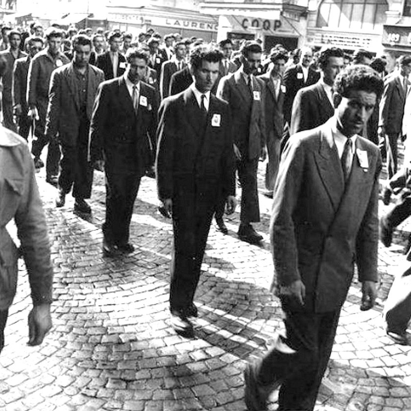 L'image montre un groupe d'hommes marchant ensemble dans une rue pavée. Tous portent des costumes et semblent sérieux. Ils avancent en rang, avec des badges sur leur veste. En arrière-plan, on peut distinguer quelques bâtiments et enseignes, ce qui donne un sens d'époque à la scène. L'atmosphère peut paraître solennelle, suggérant possiblement une manifestation ou un événement organisé.