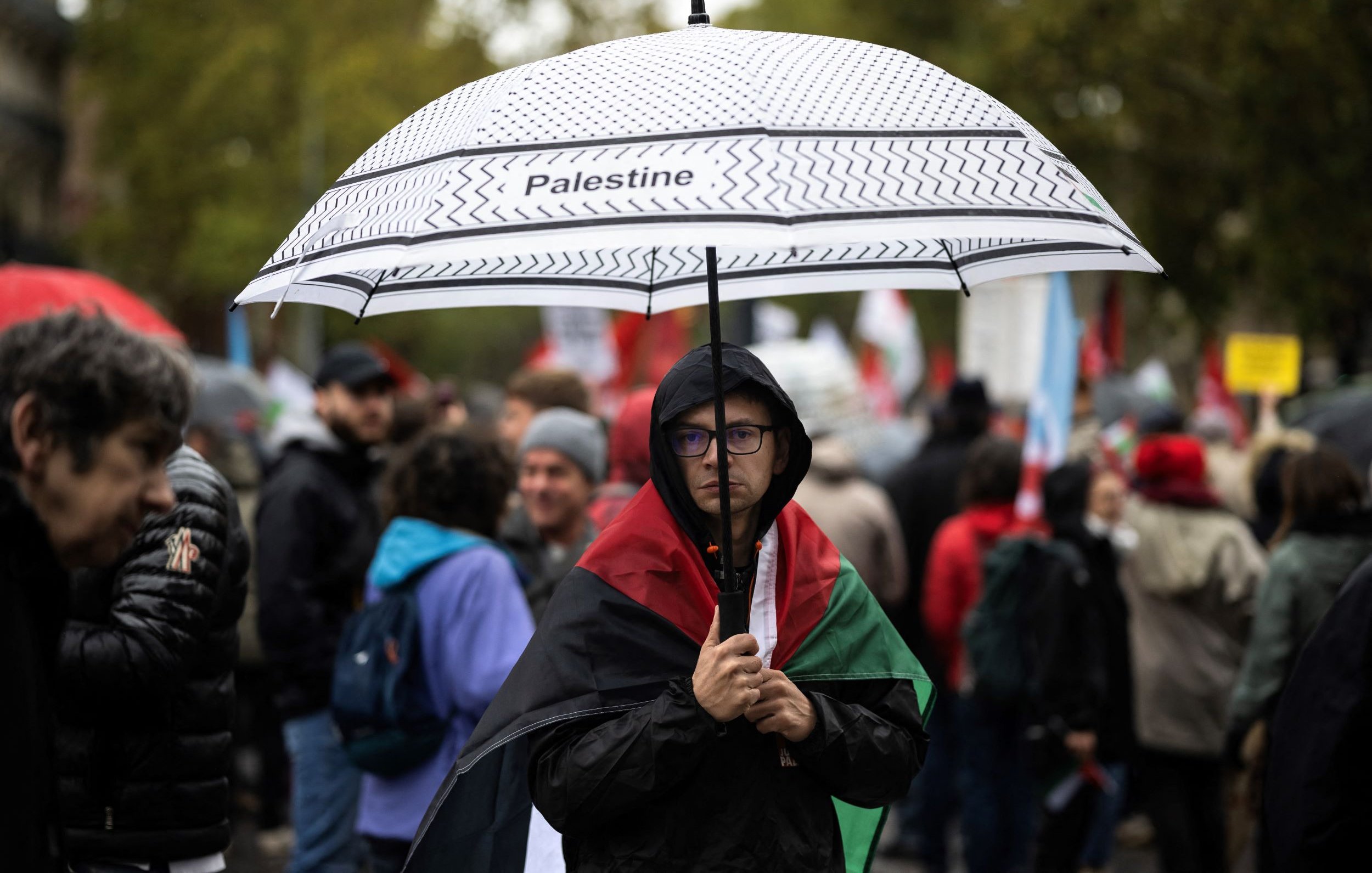 En la imagen, se puede ver a una persona que sostiene un paraguas con la palabra "Palestine" escrita en él. La persona lleva una bandera de Palestina sobre los hombros y parece estar en una manifestación o protesta. Alrededor se pueden observar otras personas, algunas con paraguas y banderas, en un ambiente cargado de emociones y solidaridad. El clima parece nublado o lluvioso, lo que añade a la atmósfera del evento.