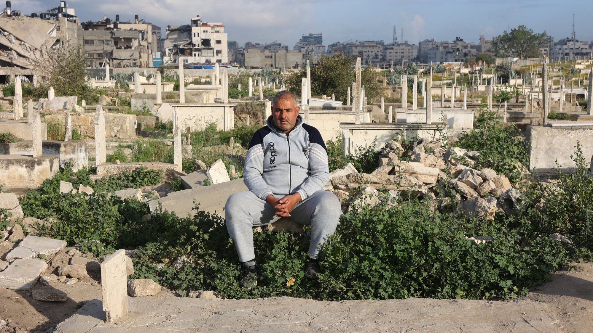 Un uomo seduto in un cimitero, circondato da tombe e vegetazione.