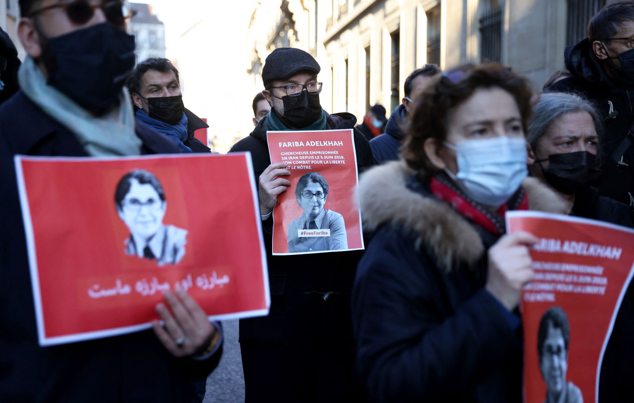 L'image montre un groupe de personnes participant à une manifestation. Elles tiennent des pancartes avec l'image et le nom d'une personne, Farida Adlénah, accompagnées de slogans. Les manifestants portent des masques, probablement en raison de préoccupations sanitaires. L'ambiance semble être solennelle et engagée, avec des visages sérieux. Les arrière-plans montrent des bâtiments qui pourraient avoir une architecture urbaine.