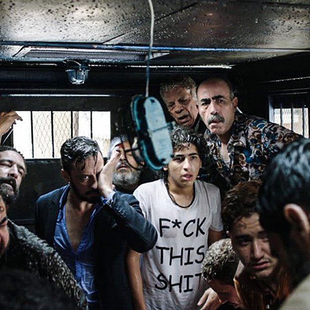 L'image montre un groupe de personnes dans un espace clos, visiblement en détresse ou en désespoir. Les visages expriment des émotions fortes, allant de la peur à l'angoisse. Certains sont habillés de manière décontractée, et l'un des individus porte un t-shirt avec un message provocateur. L'éclairage est faible, créant une ambiance sombre et oppressante. L'environnement suggère une situation de tension ou de crise.