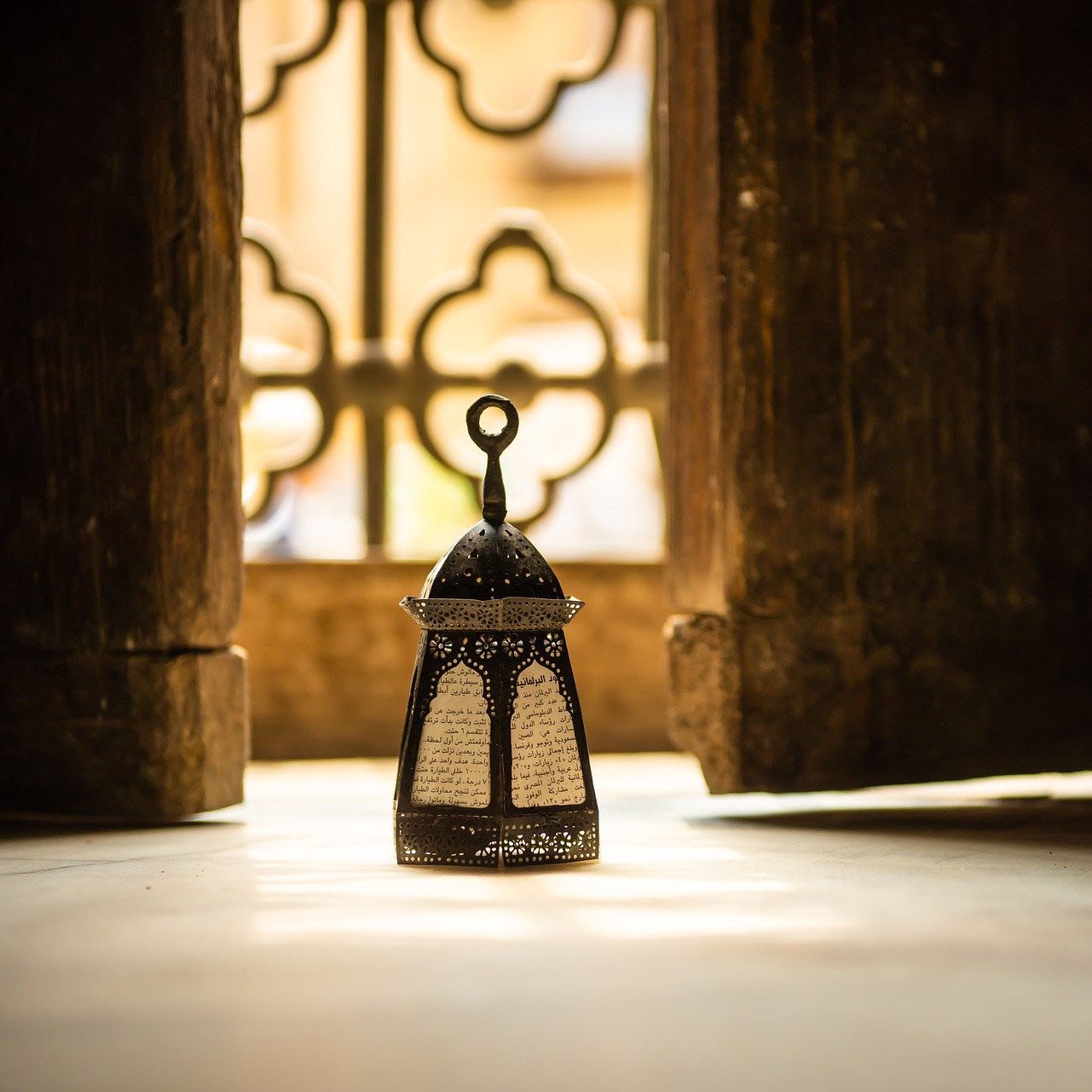 L'image montre une délicate lanterne orientale placée sur le sol, éclairée par une lumière douce, créant une atmosphère chaleureuse. En arrière-plan, on aperçoit un mur avec des motifs décoratifs, et une fenêtre aux formes ornées, laissant transparaître des lumière et ombres intéressantes. La combinaison de la lanterne et de l'environnement évoque une ambiance paisible et méditative.