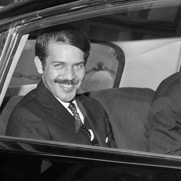 The image shows a man sitting in the back seat of a car, smiling at the camera. He is dressed in a formal suit and tie, with his hair neatly styled. Another person, partially visible, is seated next to him, also dressed in a suit. The setting appears to be a city, indicated by buildings in the background. The photograph has a classic black-and-white aesthetic, suggesting it was taken several decades ago.