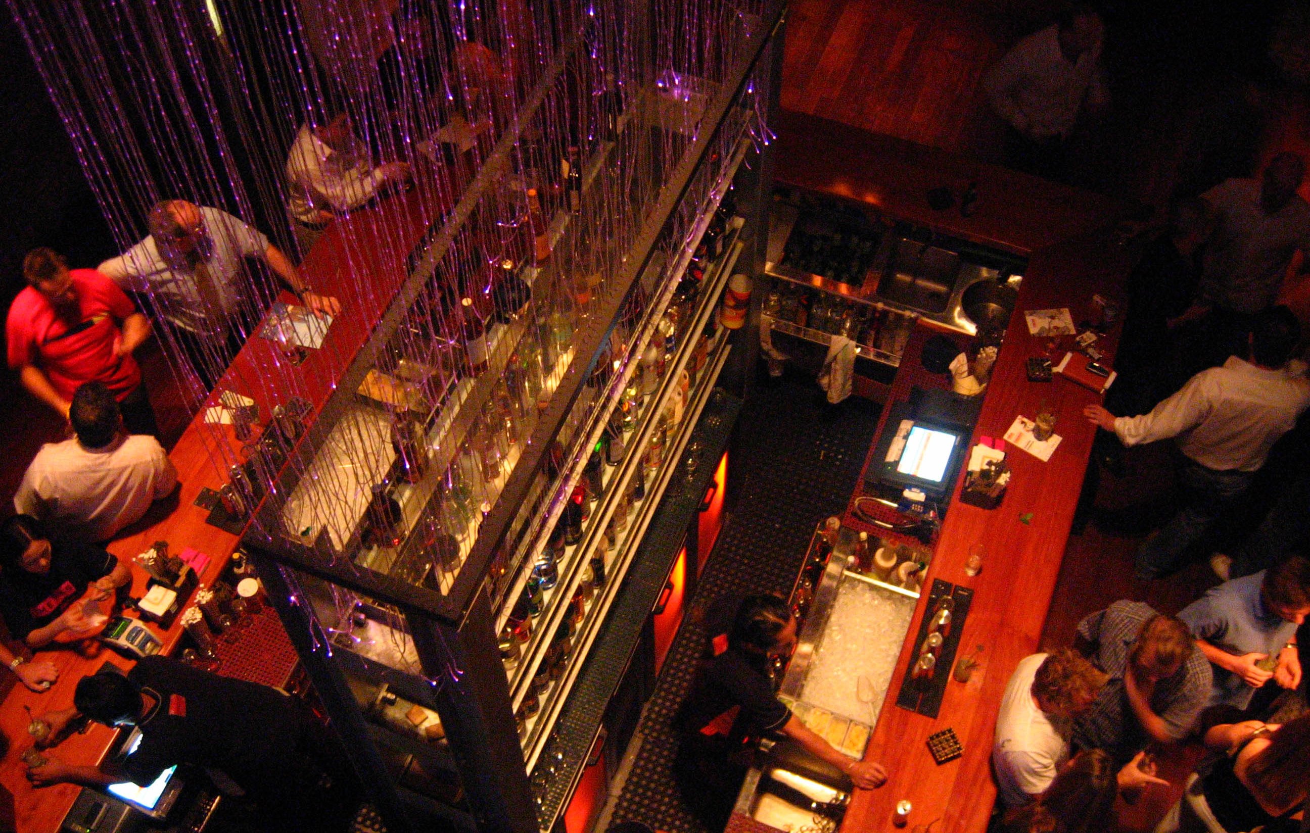 The image depicts a lively bar scene viewed from above. The bar features multiple shelves filled with bottles and is adorned with decorative lights. People are gathered throughout the space, engaging in conversation and enjoying drinks. The atmosphere seems vibrant and social, with a mix of patrons at the bar and tables. The lighting adds a warm, inviting ambiance to the setting.