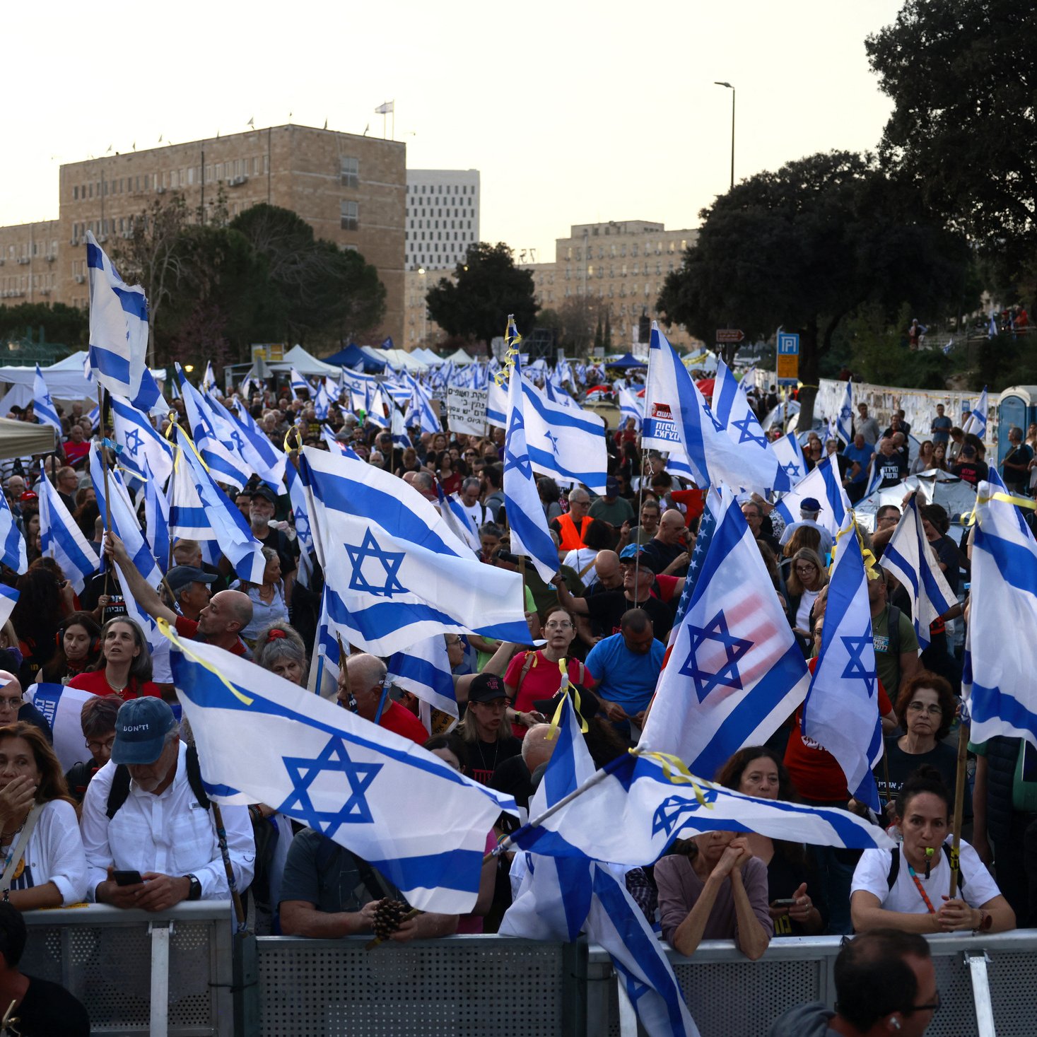 L'immagine mostra una folla di persone che partecipa a una manifestazione, sventolando molte bandiere di Israele. Il pubblico sembra essere molto numeroso e coinvolto, con alcuni partecipanti che usano telefoni cellulari. Sullo sfondo si possono vedere statue e edifici, mentre la scena è illuminata dalla luce del giorno. L'atmosfera appare vivace e carica di spirito collettivo.