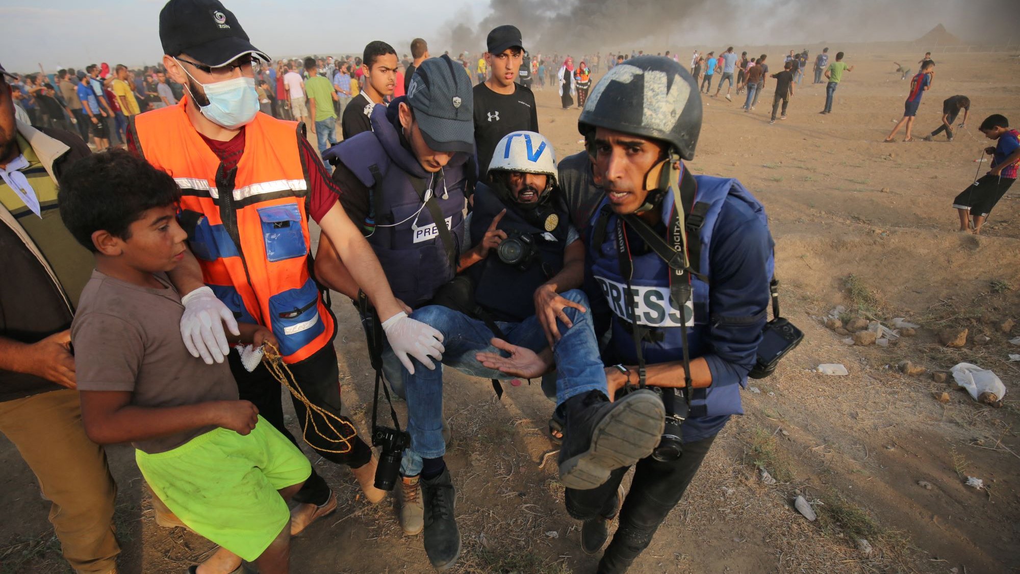 L'immagine mostra un gruppo di persone in una situazione di emergenza. Alcuni uomini, vestiti con giubbotti di protezione e tesserini "PRESS", stanno aiutando un uomo che è visibilmente ferito, portandolo via dal luogo del conflitto. Sullo sfondo si vede del fumo, suggerendo che ci sia stata un'esplosione o un incendio. Un ragazzo in primo piano osserva la scena con attenzione. L'atmosfera è tesa e drammatica, evidenziando una situazione di crisi.