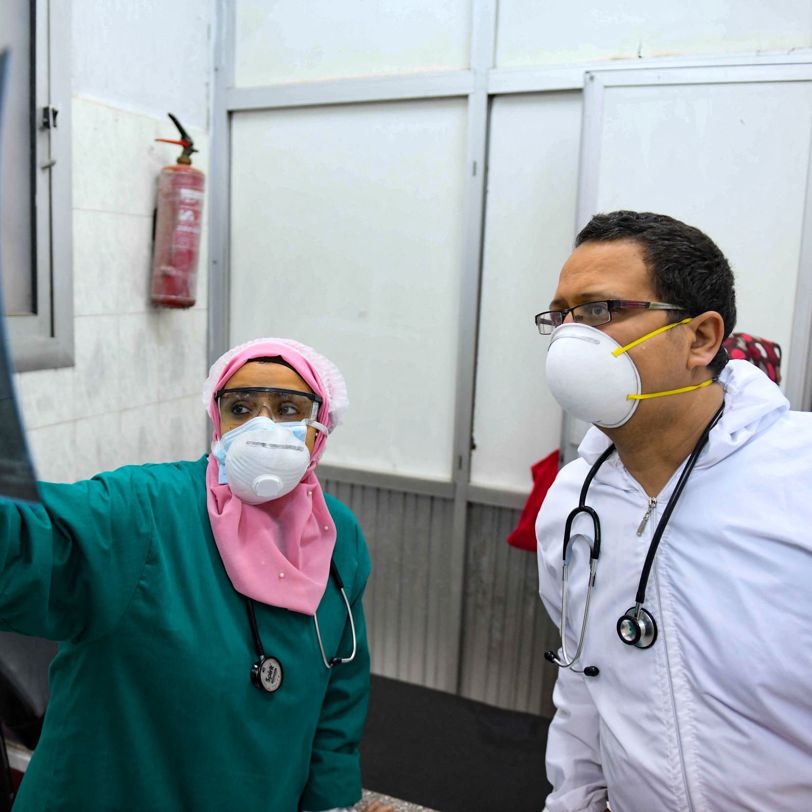 L'image montre deux professionnels de la santé examinant une radiographie. La femme porte un uniforme vert, un masque et un foulard rose, tandis que l'homme est vêtu d'une blouse blanche et porte également un masque. Ils se tiennent dans une pièce médicale, entourés de murs en carrelage. On peut voir un extincteur accroché au mur. L'expression de concentration indique qu'ils discutent sérieusement des résultats de l'examen.
