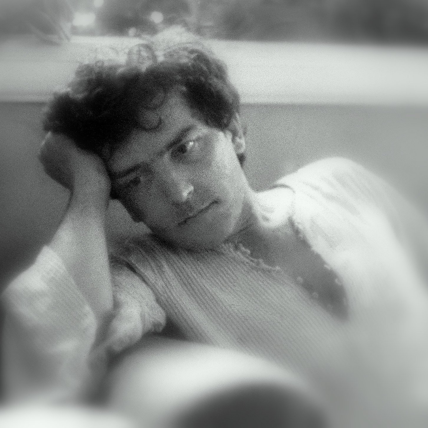 L'image montre un jeune homme pensif, appuyé sur sa main, avec une expression mélancolique. Il semble perdu dans ses pensées, ses cheveux sont légèrement ondulés et il porte une chemise à manches longues. L'arrière-plan est flou, créant une ambiance douce et introspective. La lumière est tamisée, ajoutant à l'atmosphère de réflexion.