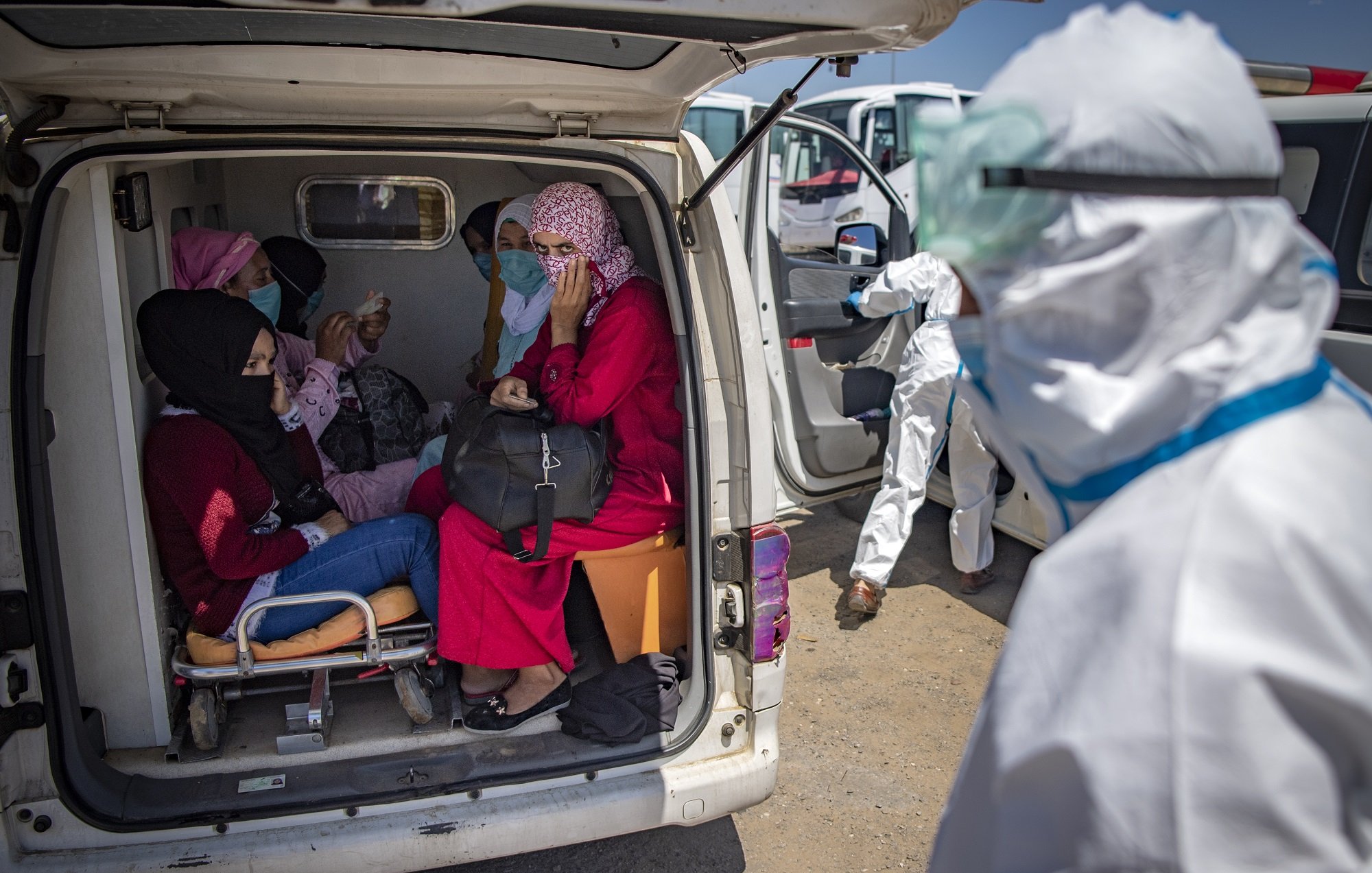 L'image montre un groupe de personnes à l'intérieur d'une ambulance. On peut voir plusieurs femmes assises, certaines portant des masques et des vêtements colorés. Une femme porte un hijab et semble discuter avec les autres. À l'extérieur, une personne en tenue de protection, probablement un professionnel de la santé, s'approche. L'environnement semble être une zone d'intervention médicale. La scène évoque une atmosphère de soin et d'assistance dans un contexte de santé.
