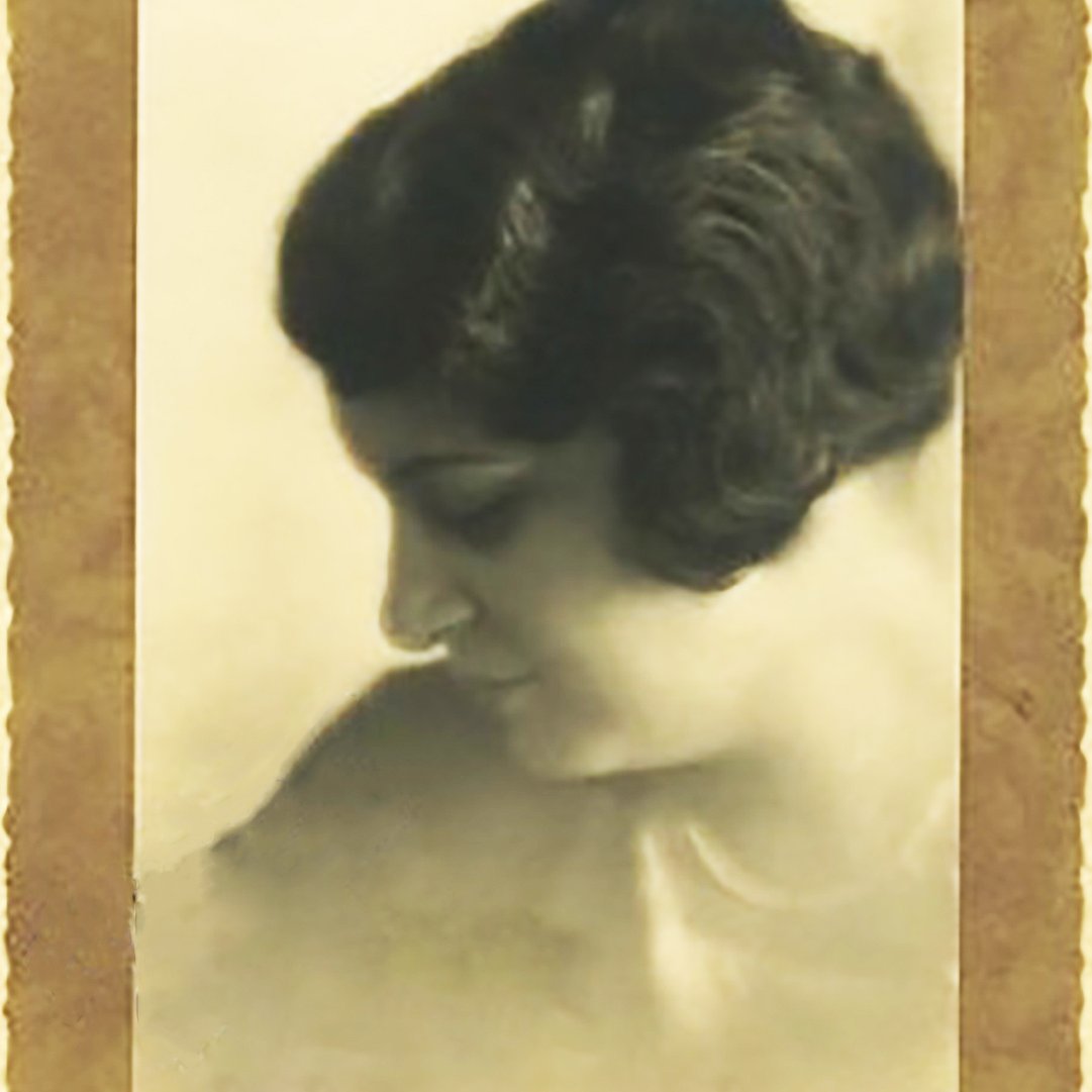 L'image représente un portrait en noir et blanc d'une femme, pris de profil. Elle a les cheveux bouclés et une coiffure élégante typique des années 1920-1930. Son expression est douce et pensive, et elle semble regarder vers le bas, créant une ambiance contemplative. Le fond de l'image est flou, mettant en valeur son visage, et le tout est encadré par une bordure décorative qui ajoute une touche vintage à la photo.