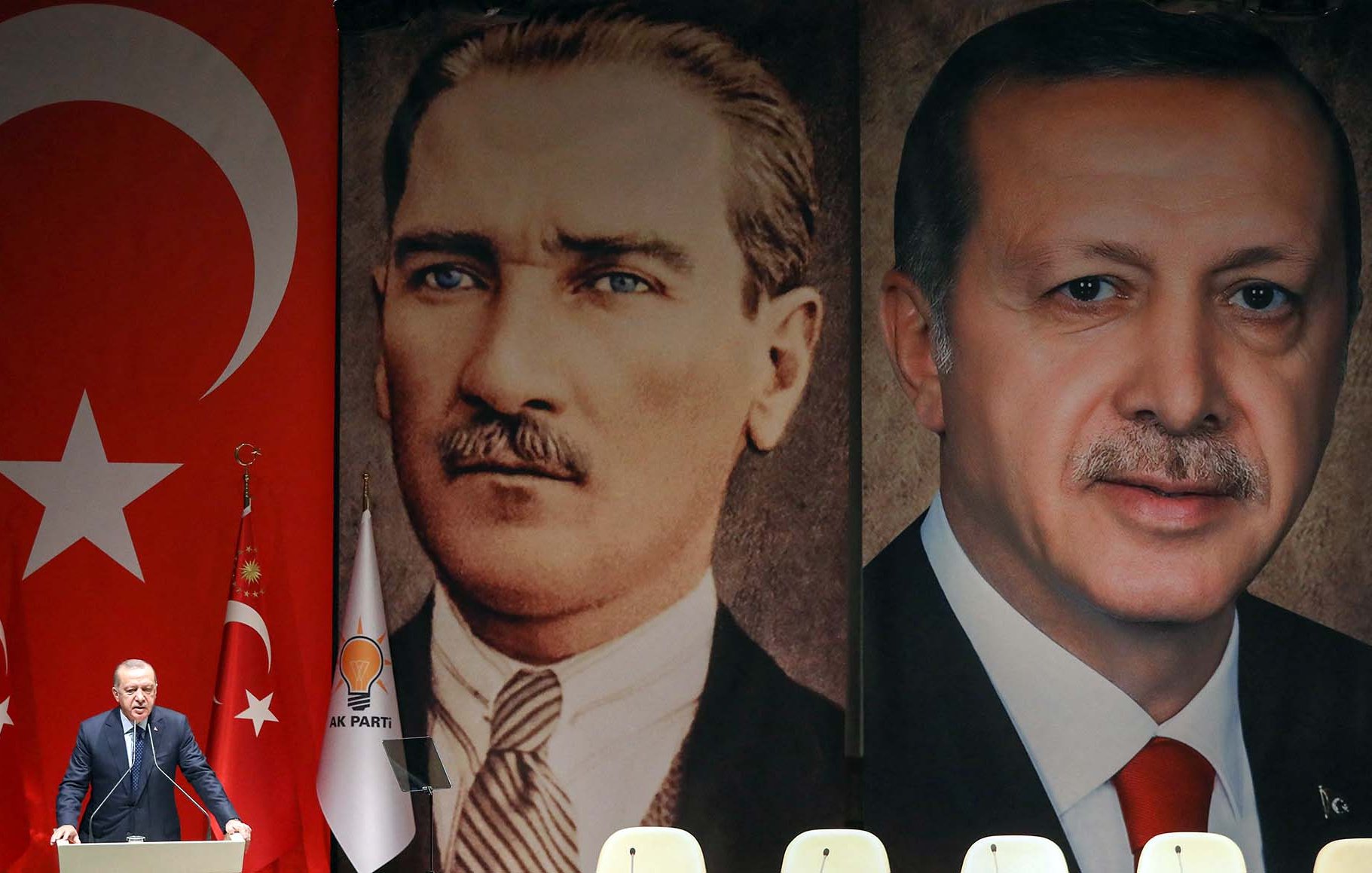 L'image présente un homme debout devant une scène décorée de grands portraits de deux figures importantes de la Turquie. À gauche, on voit le portrait de Mustafa Kemal Atatürk, le fondateur de la République turque, et à droite, celui de Recep Tayyip Erdoğan, l'actuel président. En arrière-plan, il y a un grand drapeau turc. L'homme sur la scène semble s'adresser à un public, probablement lors d'un discours politique. Des drapeaux et des emblèmes associés au parti politique d'Erdoğan sont également visibles.