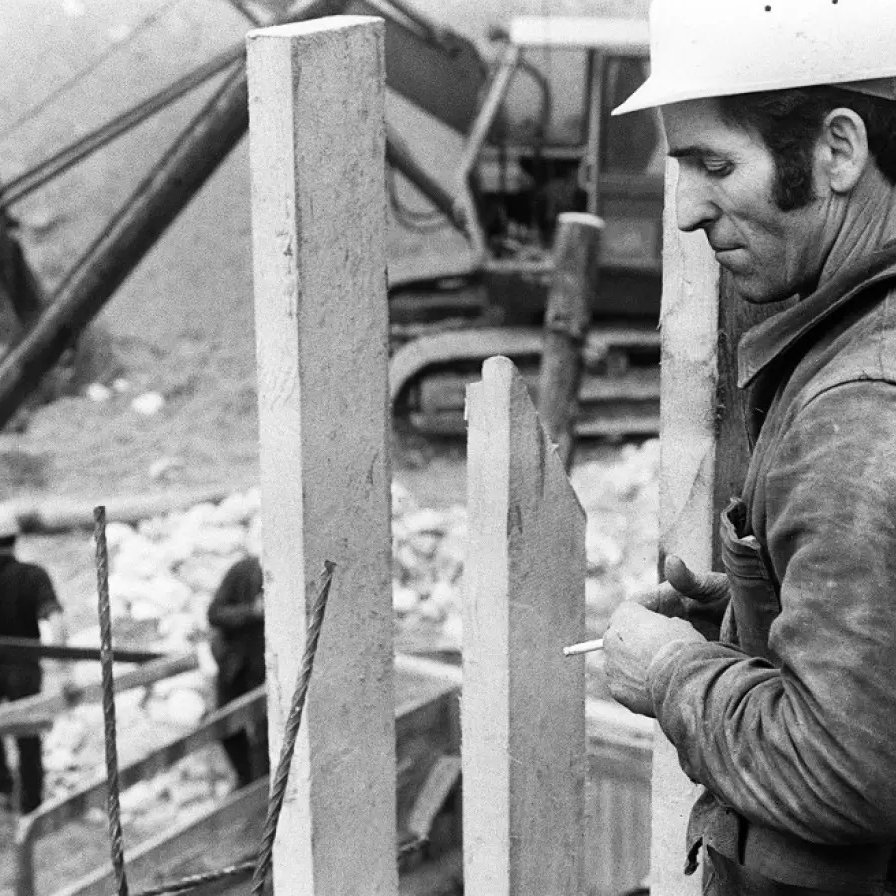 L'image montre un homme en train de travailler sur un chantier de construction. Il porte un casque de protection blanc et semble concentré sur sa tâche. À proximité, on peut voir des éléments en béton vertical, probablement des murs ou des supports. En arrière-plan, des ouvriers sont également visibles, et une machine lourde est à l'œuvre. L'image est en noir et blanc, ce qui lui donne un aspect rétro et saisissant. L'ambiance générale évoque le travail manuel et l'effort dans le domaine de la construction.