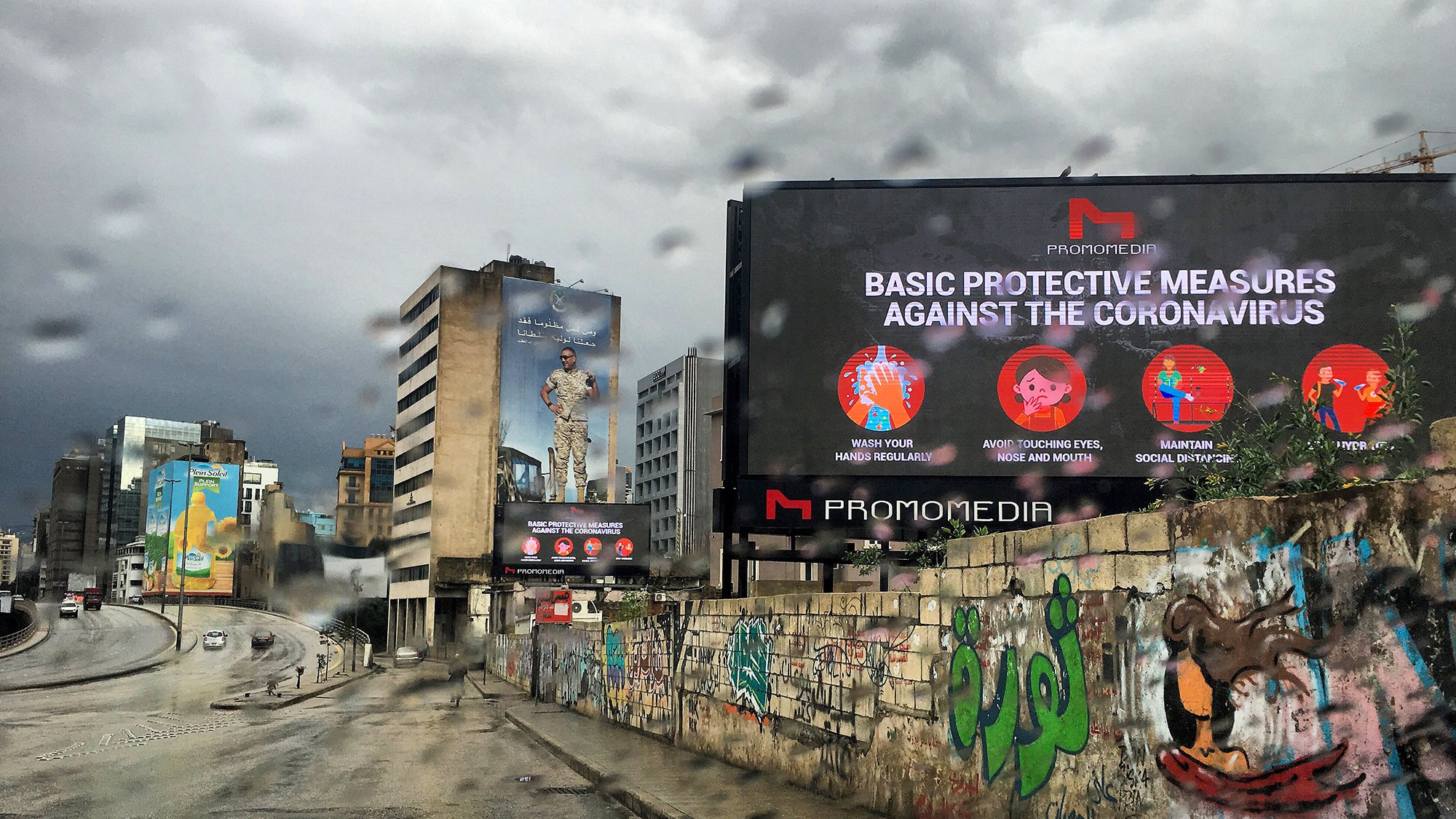 La imagen muestra una calle en un entorno urbano, con un ambiente lluvioso. Hay un letrero digital grande que presenta medidas de protección básicas contra el coronavirus. En el fondo, se pueden ver edificios y murales coloridos en las paredes, lo que añade un contraste visual a la escena. El cielo está nublado, lo que sugiere un clima gris y lluvioso.