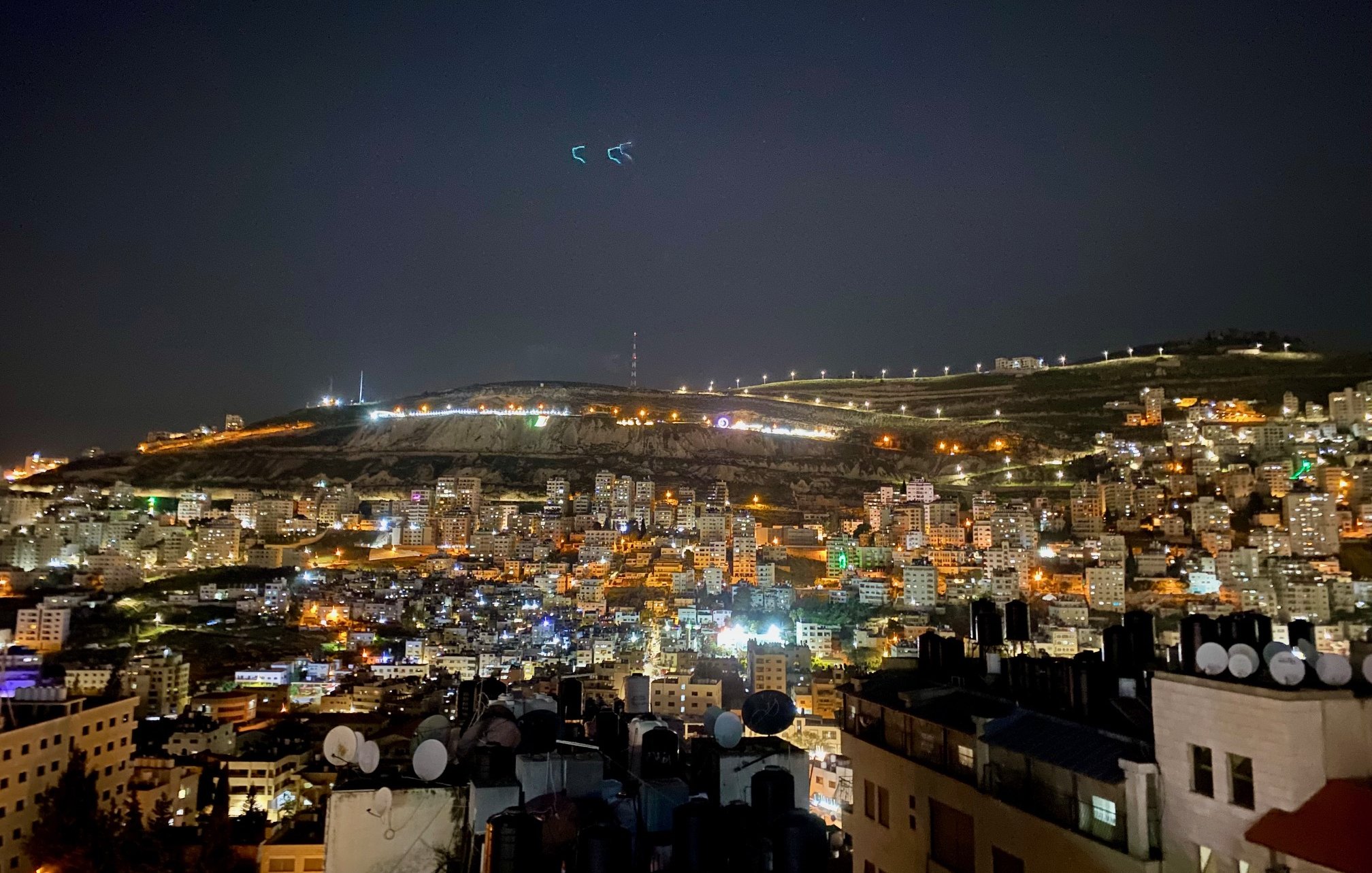 The image shows a nighttime view of a cityscape, likely on the slope of a hill or mountain. The scene is illuminated with numerous lights from buildings, giving it a vibrant and lively atmosphere. In the foreground, there are rooftops with antennas or satellite dishes visible, while the background features the illuminated hillside, possibly with telecommunications towers or other structures visible at the top. The sky is dark, suggesting it's a clear night with some stars or light reflections. Overall, the image conveys a sense of urban life and a stunning nocturnal landscape.