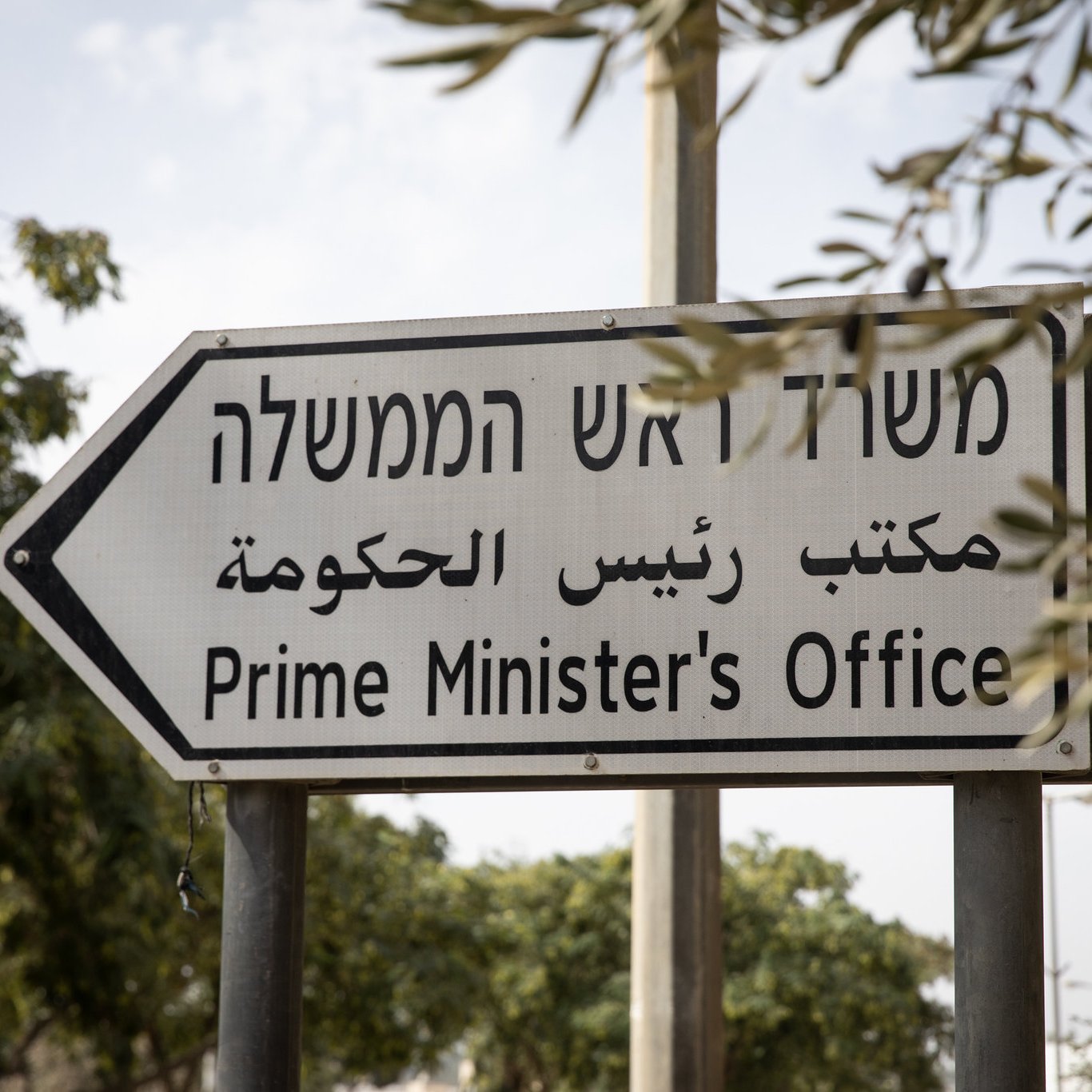 L'image montre un panneau de signalisation indiquant le "Bureau du Premier Ministre". Ce panneau est trilingue, avec des inscriptions en hébreu, en arabe et en anglais, ce qui reflète la diversité linguistique de la région. Le panneau est installé dans un environnement avec des arbres en arrière-plan.