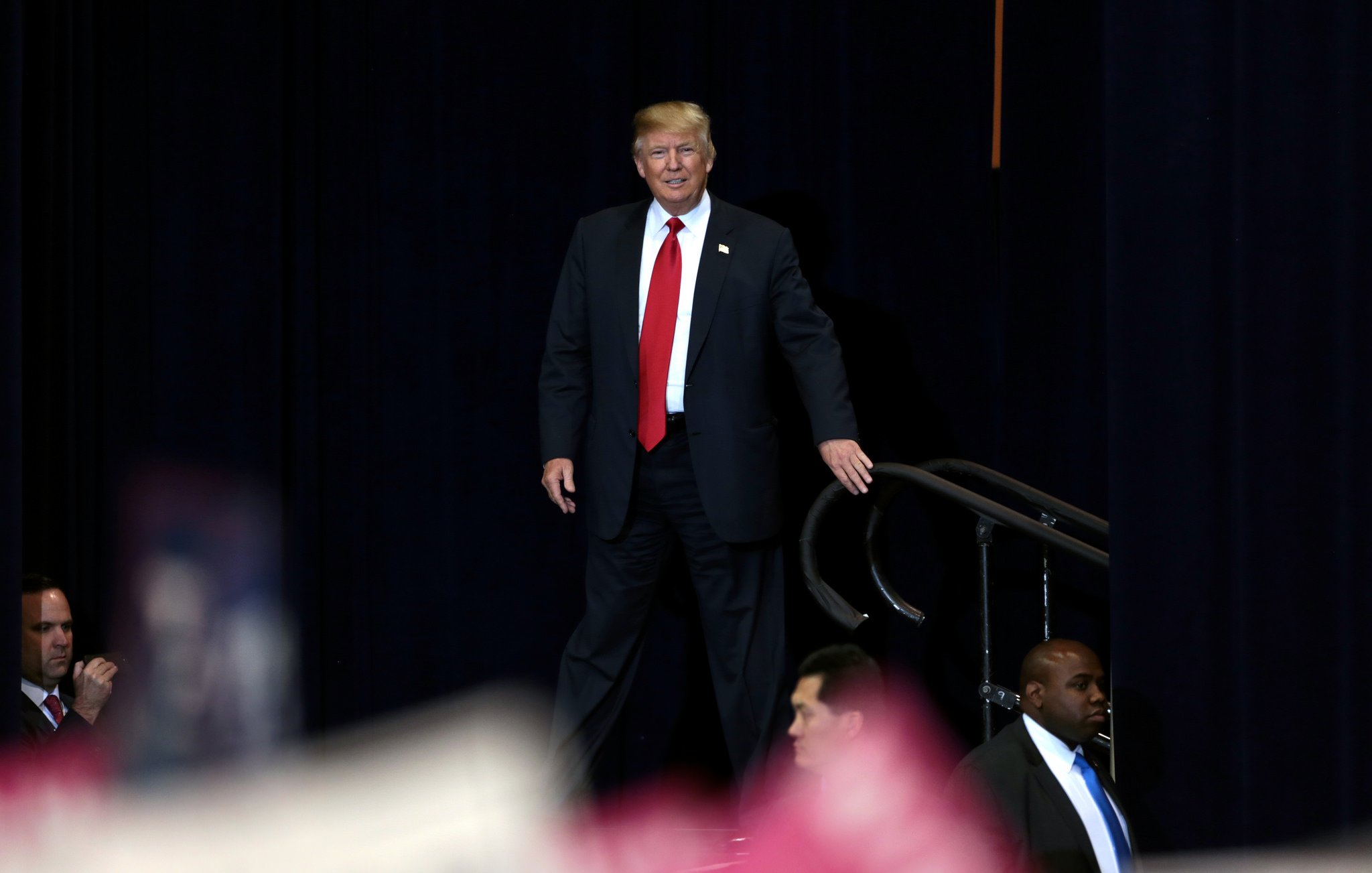 L'image montre une personne en costume, avec une cravate rouge, se tenant debout sur une scène. Il sourit et semble s'adresser à un public. Deux agents de sécurité se tiennent à proximité, surveillant la scène. L'arrière-plan est sombre, ce qui met en évidence la personne au premier plan. On voit également une foule en bas, bien que les détails soient flous.