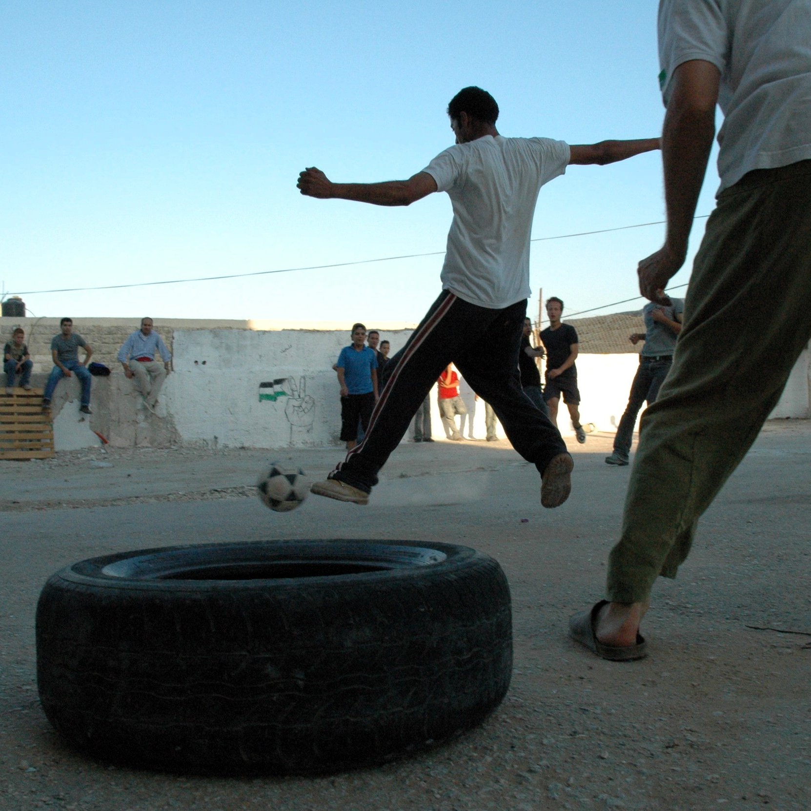 L'immagine mostra un gruppo di persone che si stanno divertendo in un ambiente all'aperto. Alcuni ragazzi stanno giocando a calcio, utilizzando un pneumatico come obiettivo. Si vedono diverse persone in piedi attorno, probabilmente che osservano la partita. La scena si svolge su un terreno sterrato, con muri bianchi sullo sfondo e un cielo azzurro. L'atmosfera è di divertimento e convivialità.