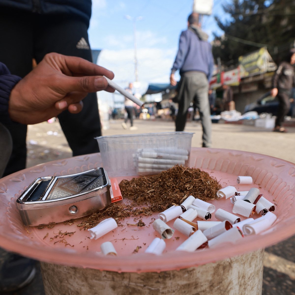L'image montre une scène de rue avec une personne qui tient une cigarette à la main. Sur un plateau, on peut voir du tabac en vrac, des cigarettes déjà consommées et un petit récipient. En arrière-plan, on aperçoit des passants et des affiches sur des murs. L'ambiance semble animée et urbaine.