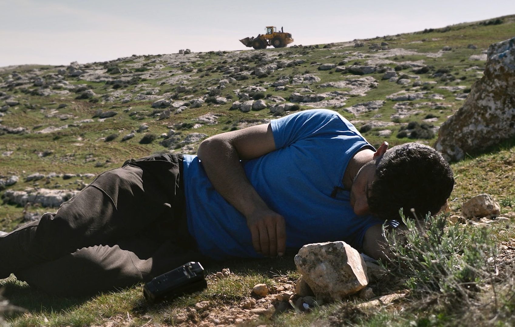 Nell'immagine si vede un uomo sdraiato su un prato verde, con il viso rivolto verso il terreno. Indossa una maglietta blu e pantaloni scuri. Accanto a lui ci sono alcune rocce. Sullo sfondo, si scorge un bulldozer che lavora su un terreno collinare. La scena trasmette un senso di tranquillità, ma anche di inquietudine a causa della presenza del macchinario che sottolinea il contrasto tra l'uomo e l'ambiente circostante.