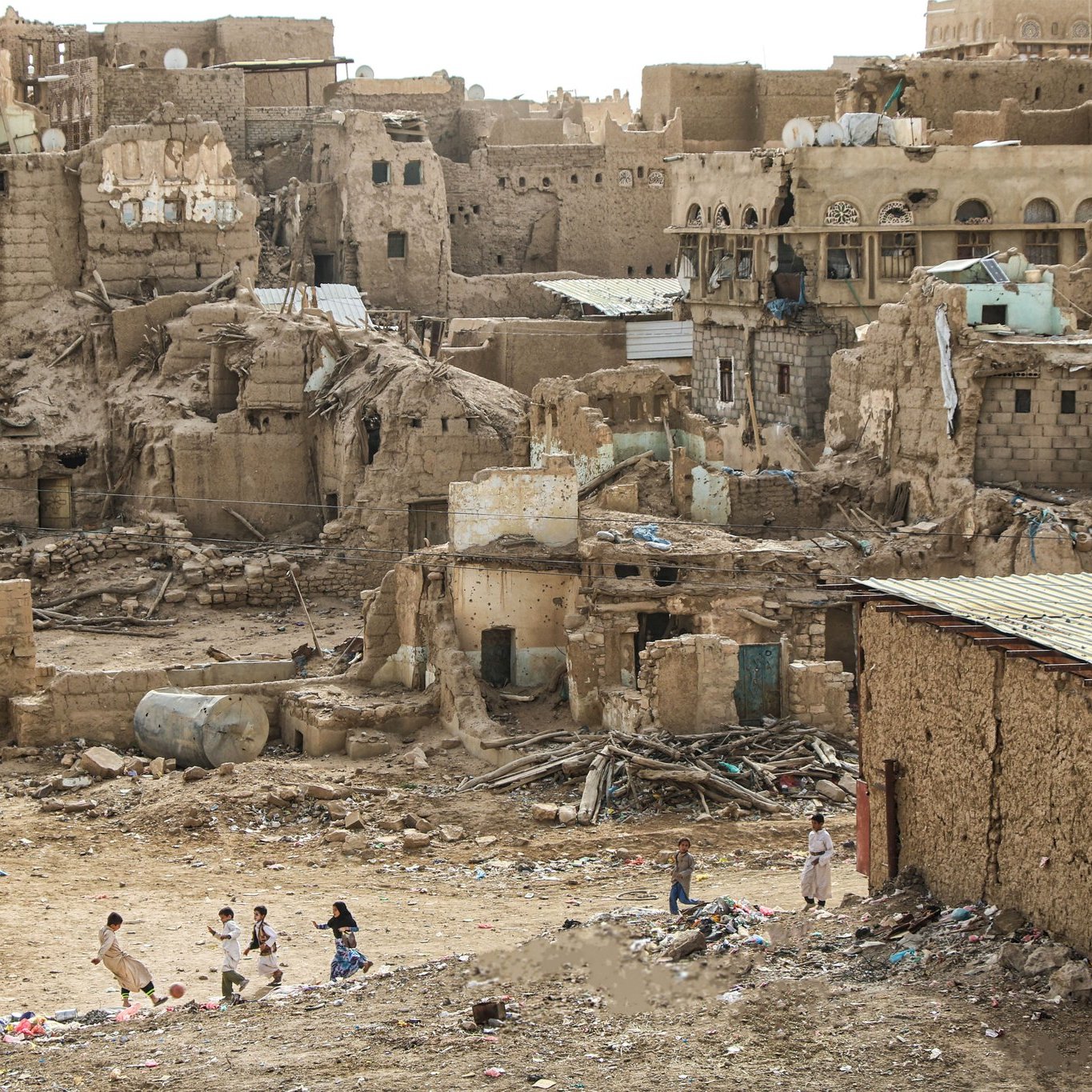 L'image montre un paysage urbain en ruines, typique d'une zone désertique. On peut voir des bâtiments en terre et en briques, avec des murs partiellement effondrés. Les structures semblent abandonnées et en mauvais état, certaines présentant des ouvertures et des décombres. Au premier plan, plusieurs enfants jouent, ce qui apporte une touche de vie à ce décor désertique. Le sol est jonché de déchets, et le ciel est clair, ce qui accentue l'aspect aride de l'environnement.