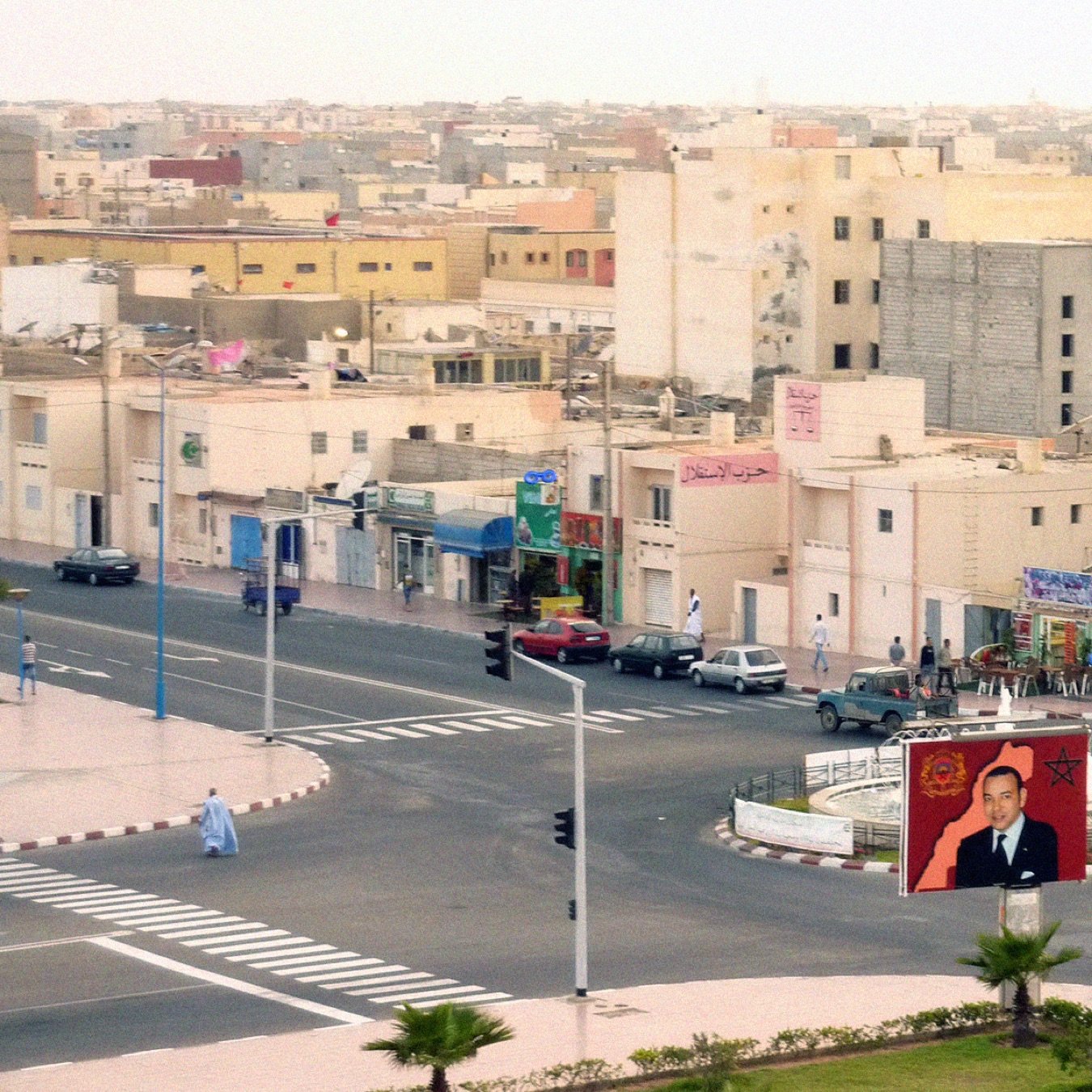 L'image montre une vue urbaine d'une ville. On y aperçoit des bâtiments de plusieurs étages, avec une architecture simple. À l'avant, il y a un rond-point avec un grand panneau affichant un portrait d'une figure publique. Plusieurs voitures sont stationnées ou circulent autour du rond-point. Des arbres et des éléments de signalisation routière ajoutent à l'ambiance de la scène. Le ciel est clair, suggérant une atmosphère calme, et la lumière semble indiquer une période de la journée avancée.