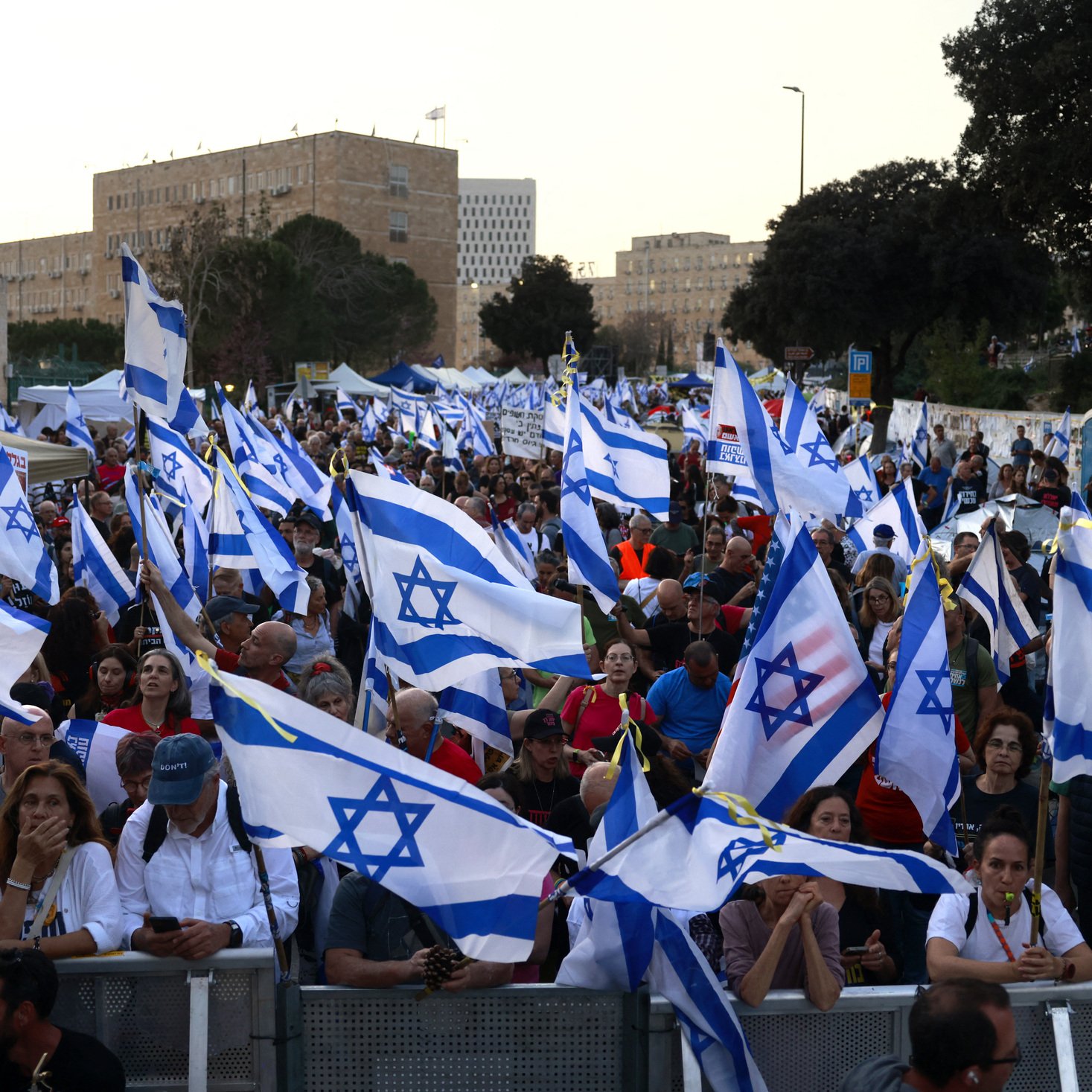The image shows a large crowd of people gathered together, holding numerous Israeli flags. The atmosphere appears to be part of a demonstration or rally, with many individuals engaged in conversation or focused on the event. The background features buildings and trees, indicating an urban setting. The flags are prominently displayed, creating a sense of unity among the participants.