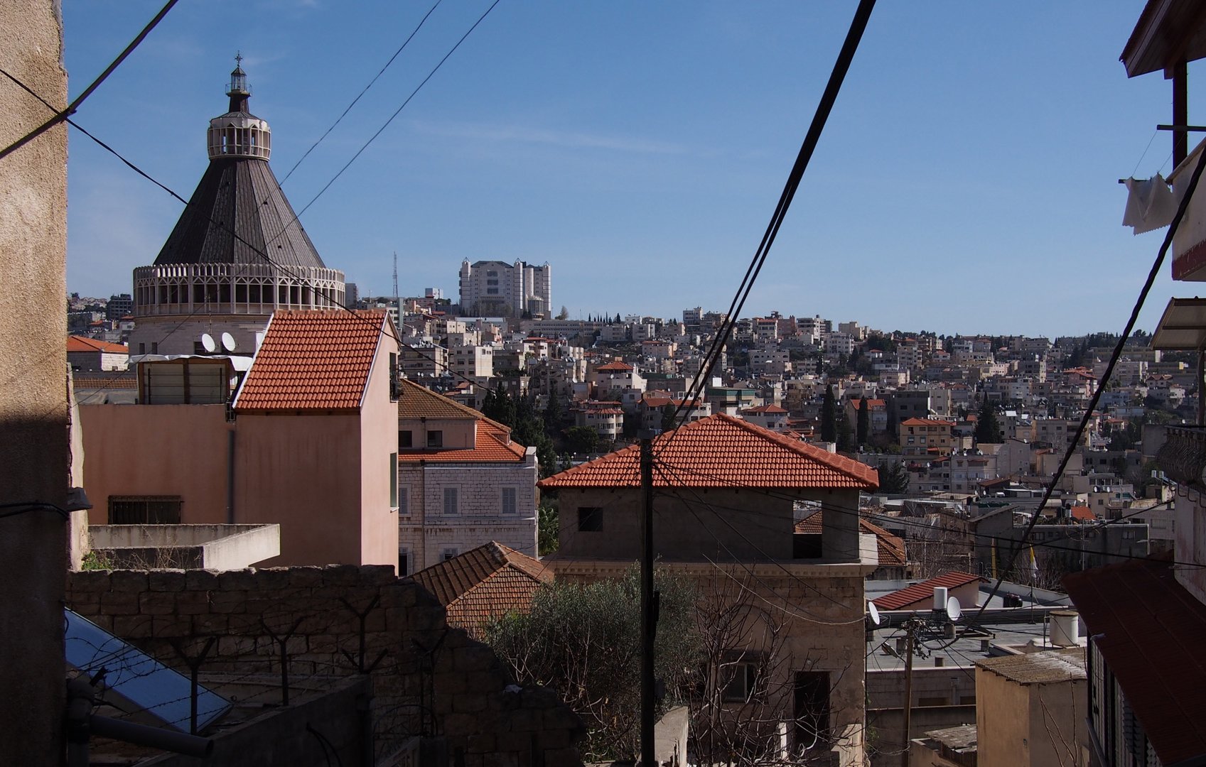 L'immagine mostra un paesaggio urbano con diverse abitazioni disposte su una collina. In primo piano, ci sono edifici con tetti a spiovente di colore rosso e strutture in muratura. Sullo sfondo si riconosce un grande edificio con una copertura a cupola, che si erge tra le case. Il cielo è sereno e limpido, contribuendo a dare un'impressione di vivacità al panorama cittadino. Inoltre, alcuni fili elettrici attraversano l'immagine, evidenziando l'aspetto urbano della scena.
