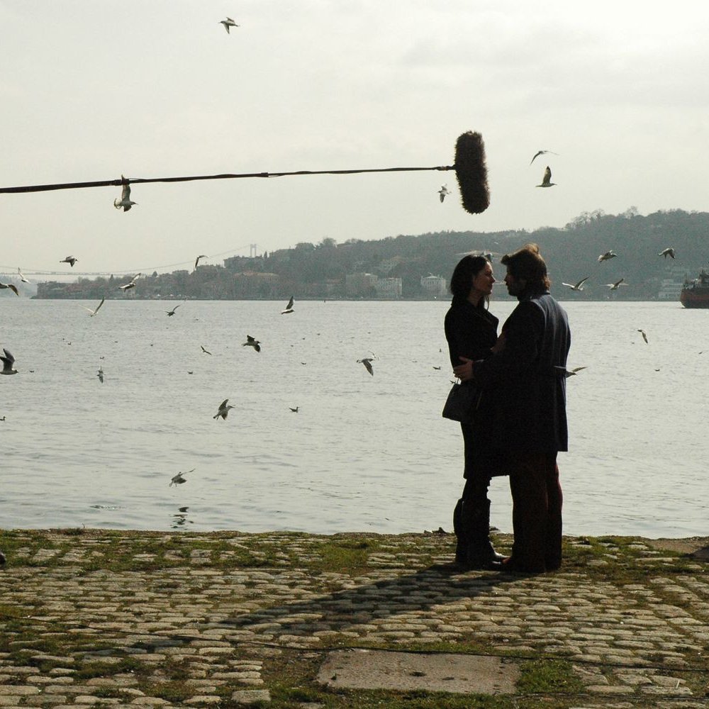 L'image montre une scène de tournage près de l'eau. On peut voir un homme tenant un perche avec un micro au-dessus de deux personnes qui semblent discuter. Ces personnes sont au premier plan, tandis qu'à l'arrière-plan, on aperçoit des oiseaux volant autour et un paysage flou. Un autre homme, au loin, semble superviser le tournage. Le temps paraît légèrement nuageux, ajoutant une atmosphère particulière à la scène.