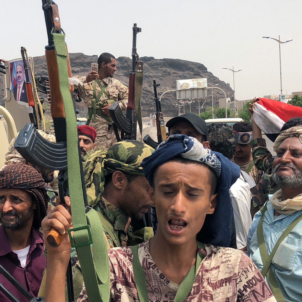 L'image représente un groupe de personnes armées, apparemment réunies dans un contexte de lutte ou de manifestation. Ils portent des vêtements typiques et semblent déterminés, avec des regards engagés. En arrière-plan, on peut apercevoir des montagnes et une circulation modérée, ainsi qu'un drapeau. Un véhicule est également visible, avec des armes visibles entre les membres du groupe. L'atmosphère suggère une situation de tension ou de conflit.