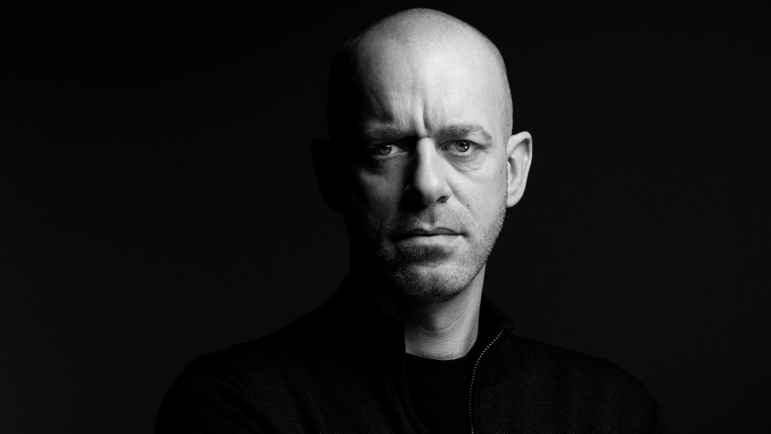 La imagen muestra a un hombre con cabeza rapada y una expresión seria. Está vestido con una prenda de color oscuro y parece estar mirando hacia la cámara. El fondo es negro, lo que resalta su figura y la iluminación hace que su rostro y gestos sean más definidos, creando un contraste dramático. La atmósfera de la imagen es intensa y poderosa.